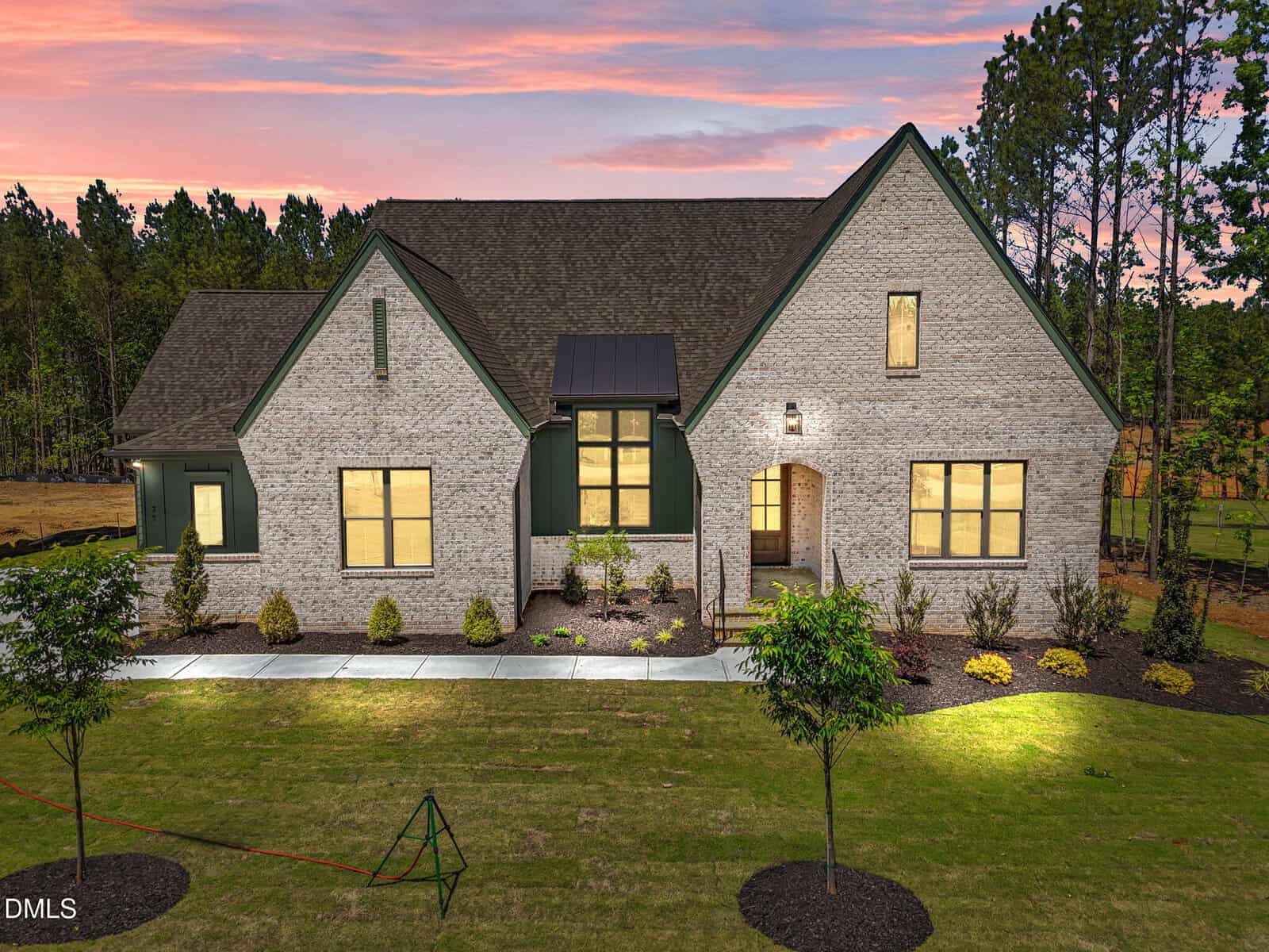 Modern two-story brick house at 35 Spanish Oak Drive, Youngsville, with large windows, green trim, and a neatly landscaped front yard, illuminated at dusk with trees and a pink-orange sunset sky in the background.