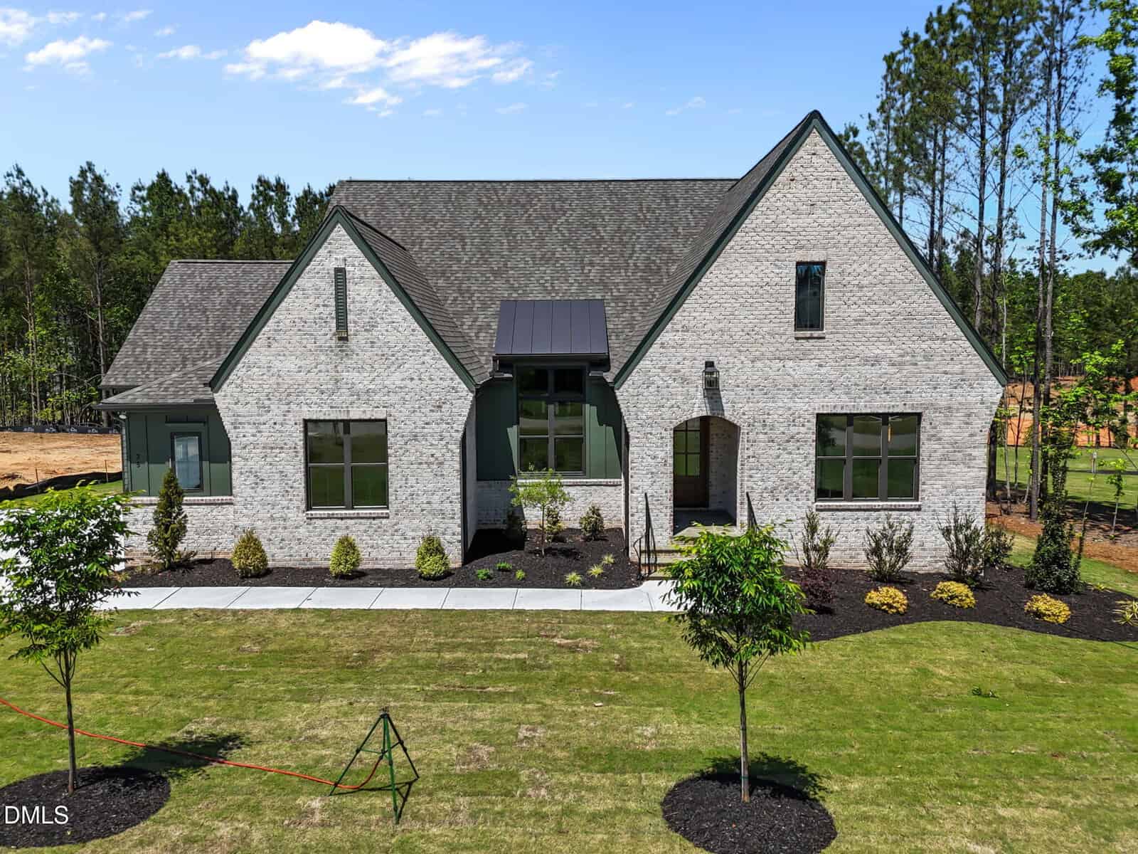 A modern, light brick house with a dark gray roof, large windows, and green trim, set on a grassy lawn with young landscaping and surrounded by trees under a partly cloudy sky.