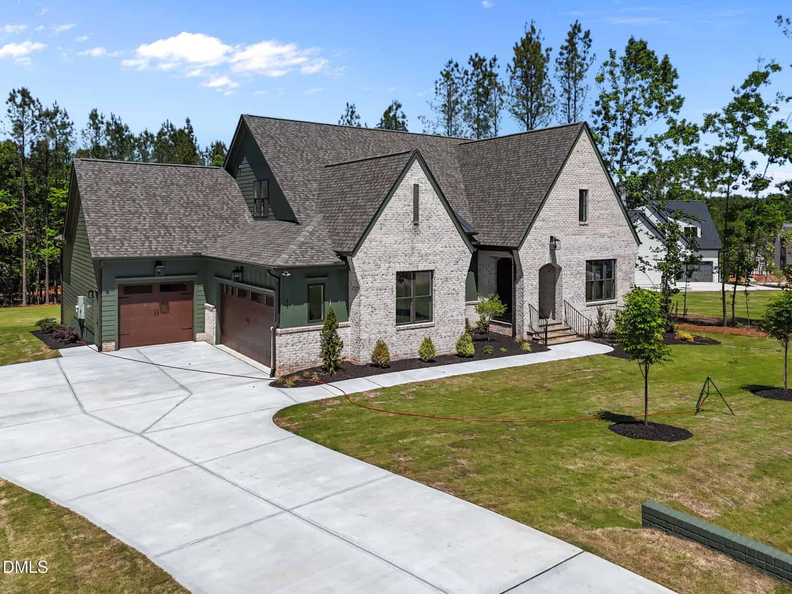 A modern two-story house with light brick exterior, green siding, and two brown garage doors, surrounded by a large lawn, young trees, and a wide driveway in a suburban neighborhood.