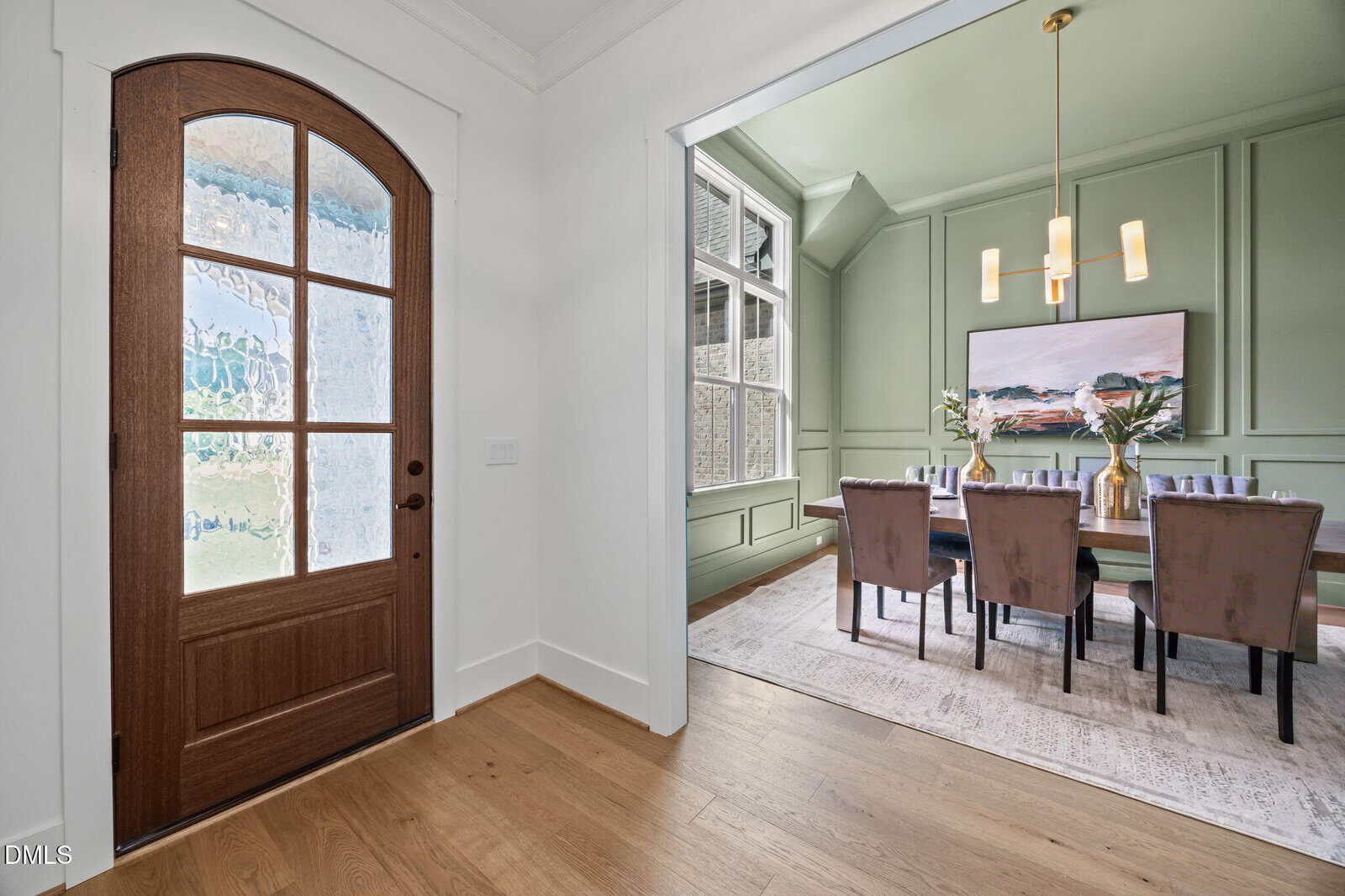 A wooden front door with glass panels opens into a home with light-colored walls and wood flooring. To the right, a dining room with green walls features a large table, upholstered chairs, a rug, and modern chandelier.