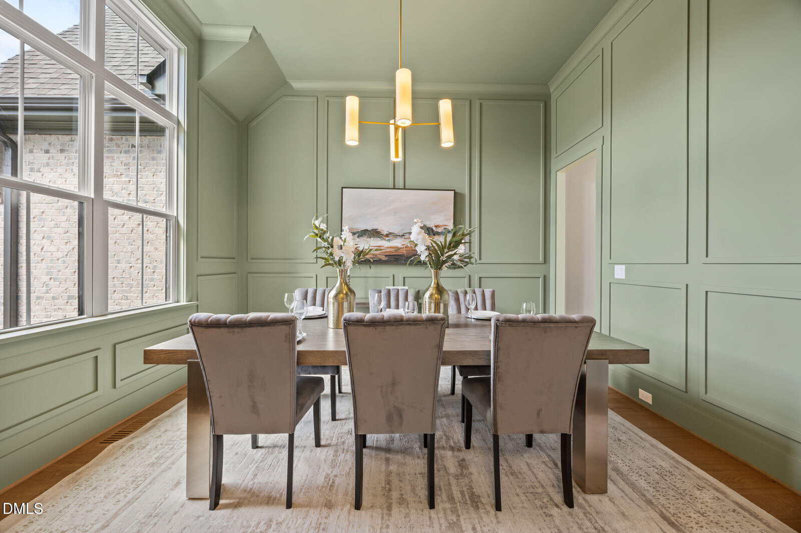 A modern dining room with sage green paneled walls, a wooden table set for six with gray upholstered chairs, floral centerpieces, an abstract painting, a gold chandelier, large windows, and a neutral rug.