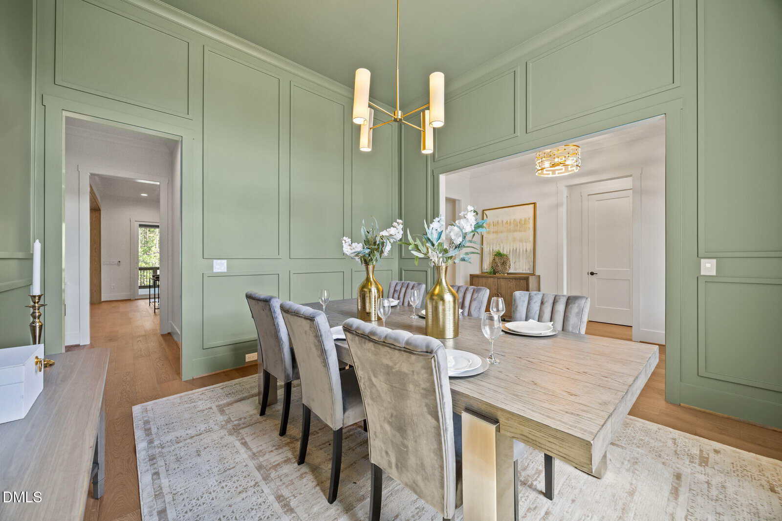 Elegant dining room with sage green paneled walls, a wooden table set for six, plush gray chairs, gold vases with flowers, and a modern chandelier. Natural light streams in from adjacent rooms.