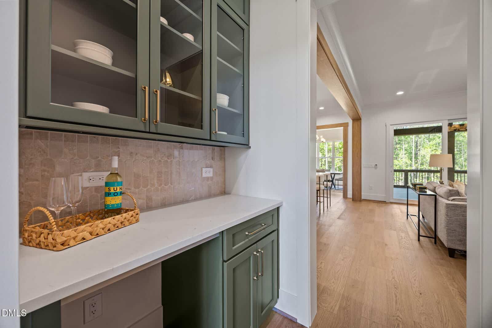 A small kitchenette features green cabinets, a white countertop with a wicker tray holding a bottle and glasses, and leads to a bright living area with large windows and a view of trees outside.