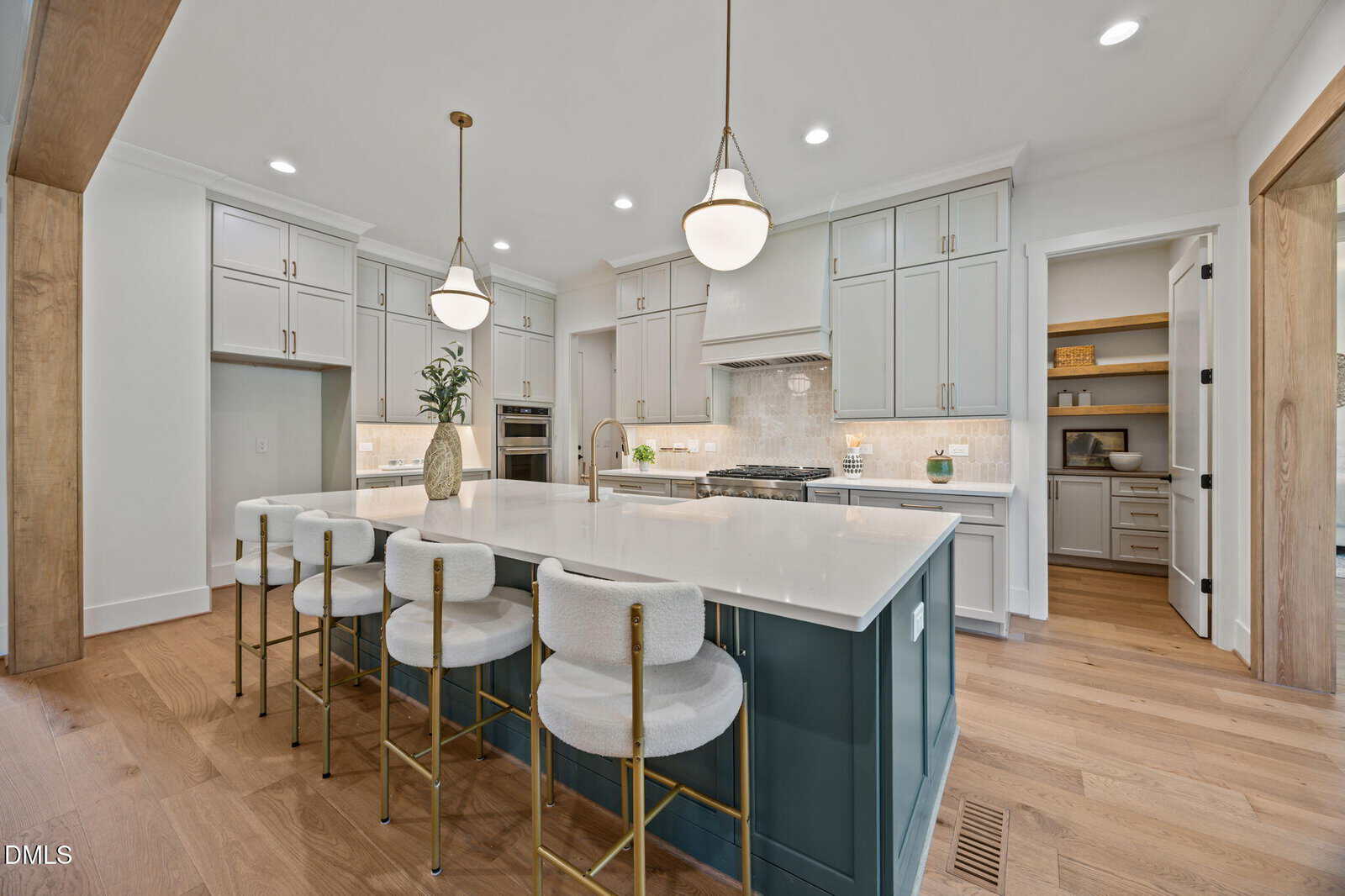 Modern kitchen with a large island, four cushioned stools, white cabinets, stainless steel appliances, pendant lights, wooden floors, and a walk-in pantry with shelves visible in the background.