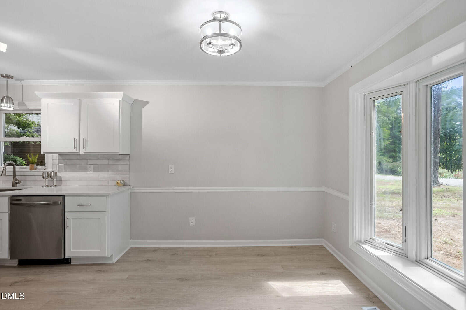 Bright kitchen and dining area with light wood floors, white cabinets, stainless steel appliances, large windows, and neutral walls. Natural light streams in, highlighting the clean and modern design.
