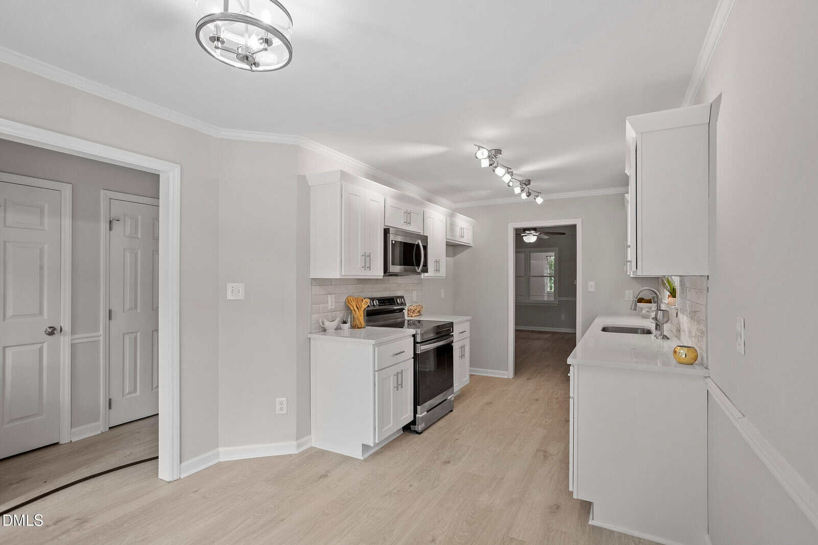 Modern kitchen with white cabinets, stainless steel appliances, light wood flooring, subway tile backsplash, and bright ceiling lights. A hallway and two closed doors are visible to the left.