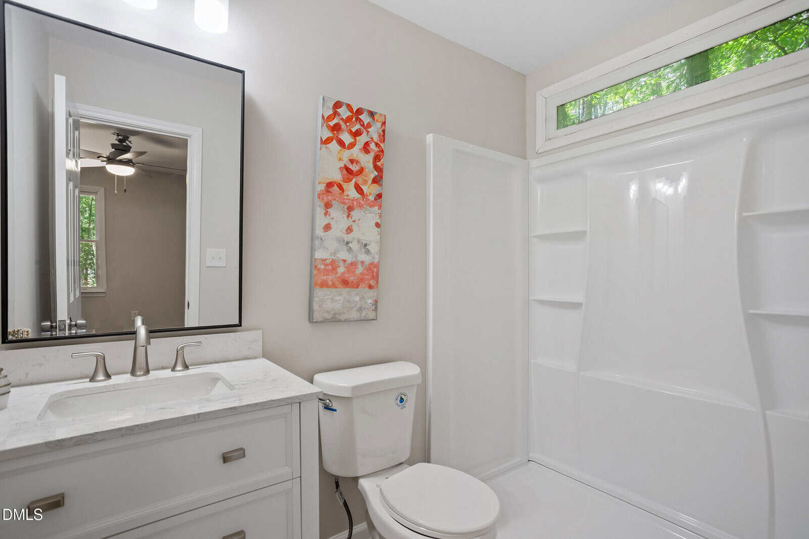 Modern bathroom with a white vanity, marble countertop, wall mirror, toilet, walk-in shower with built-in shelves, abstract red and white wall art, and a high window letting in natural light.