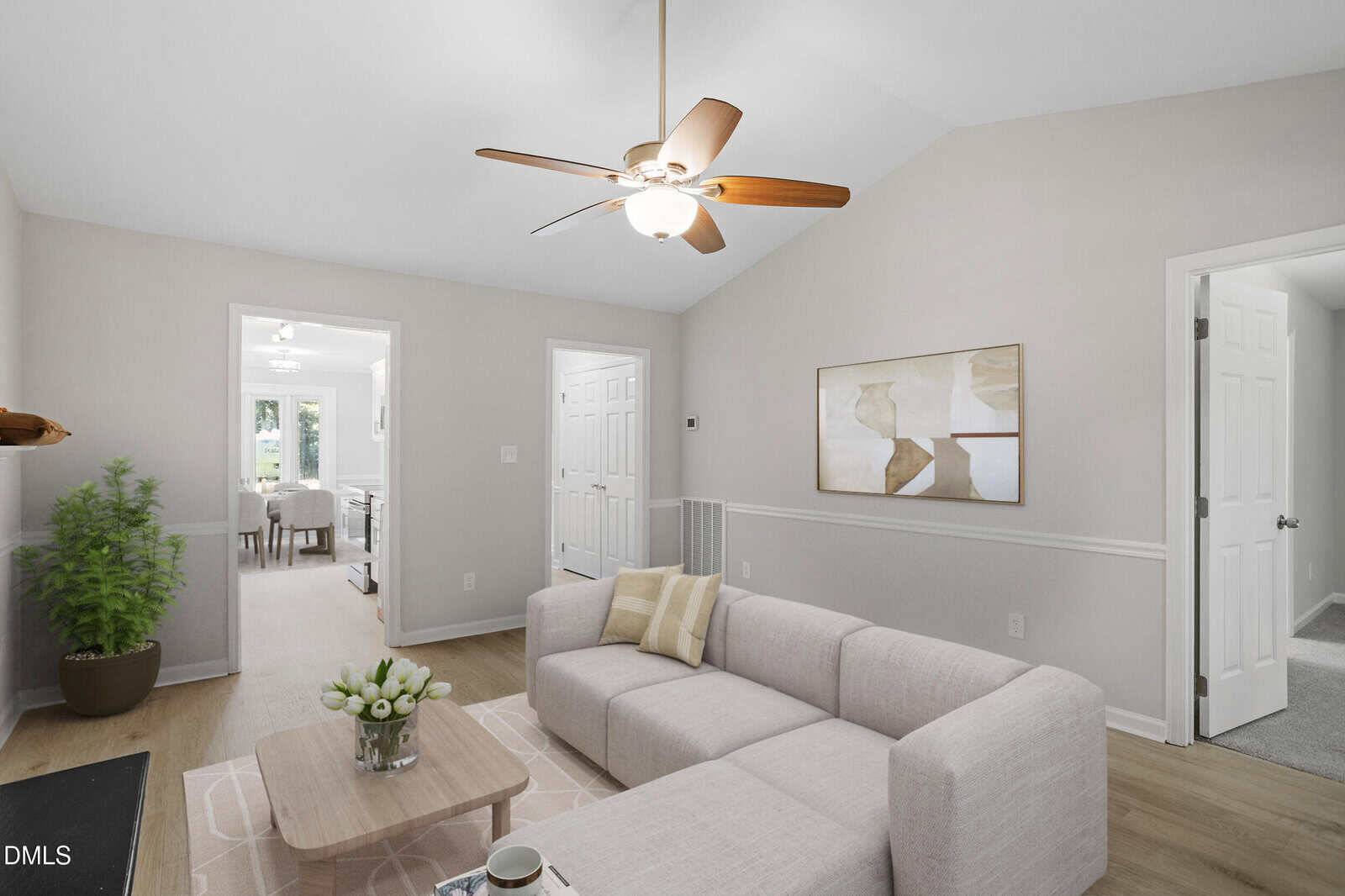 A bright living room with light walls and wood floors features a beige sectional sofa, a small coffee table with flowers, a modern ceiling fan, abstract wall art, and green plant. Doorways lead to other rooms.