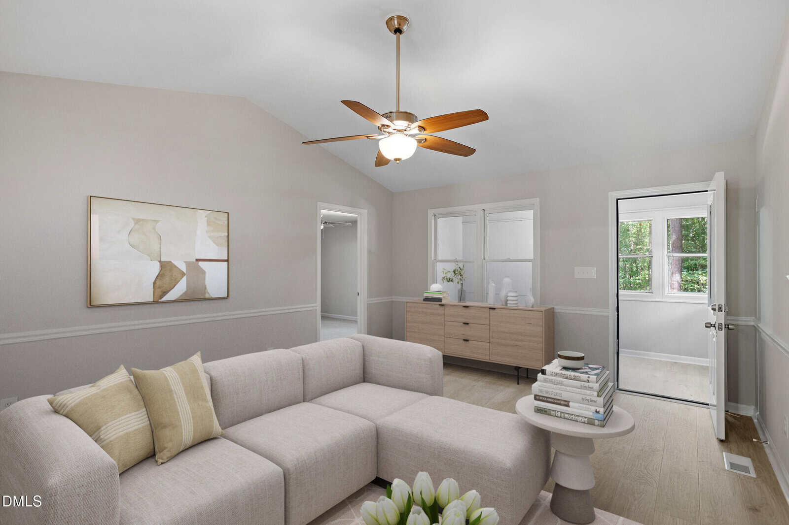 A bright living room with a beige sectional sofa, light wood floors, a round side table, minimalist artwork on the wall, a ceiling fan, and a windowed door leading outside.