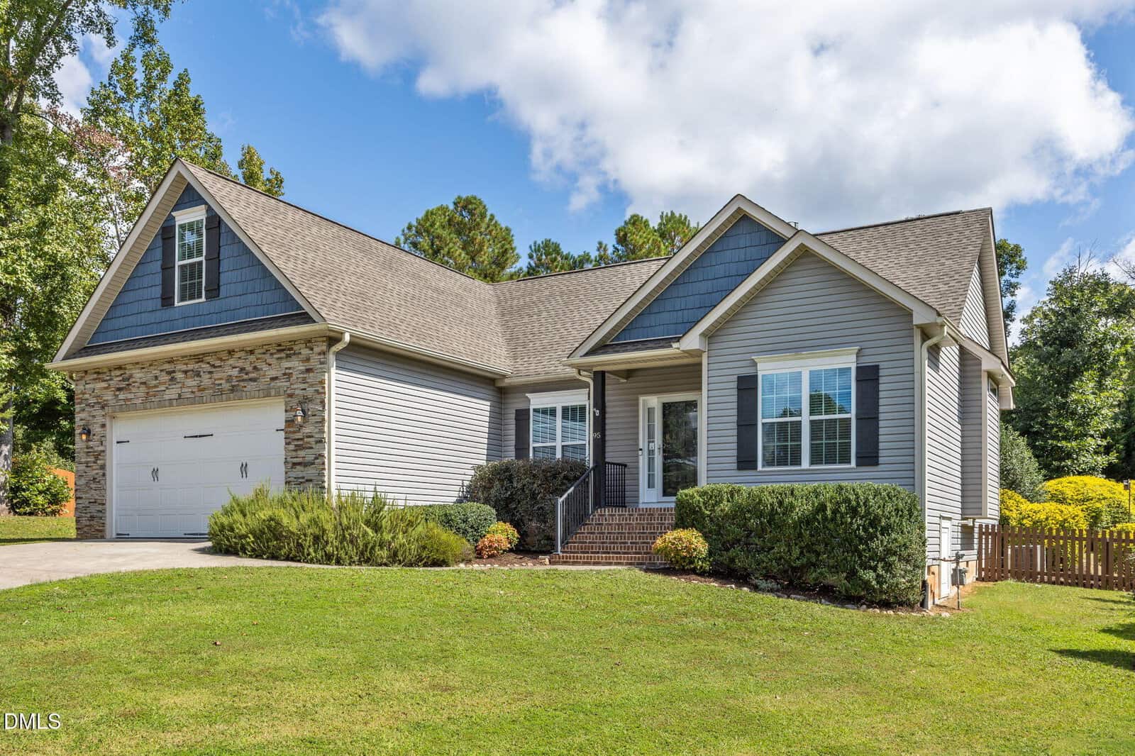 Single-story suburban house with gray siding and stone accents, attached two-car garage, front porch with steps, manicured lawn, bushes, and trees in the background under a blue sky with scattered clouds.