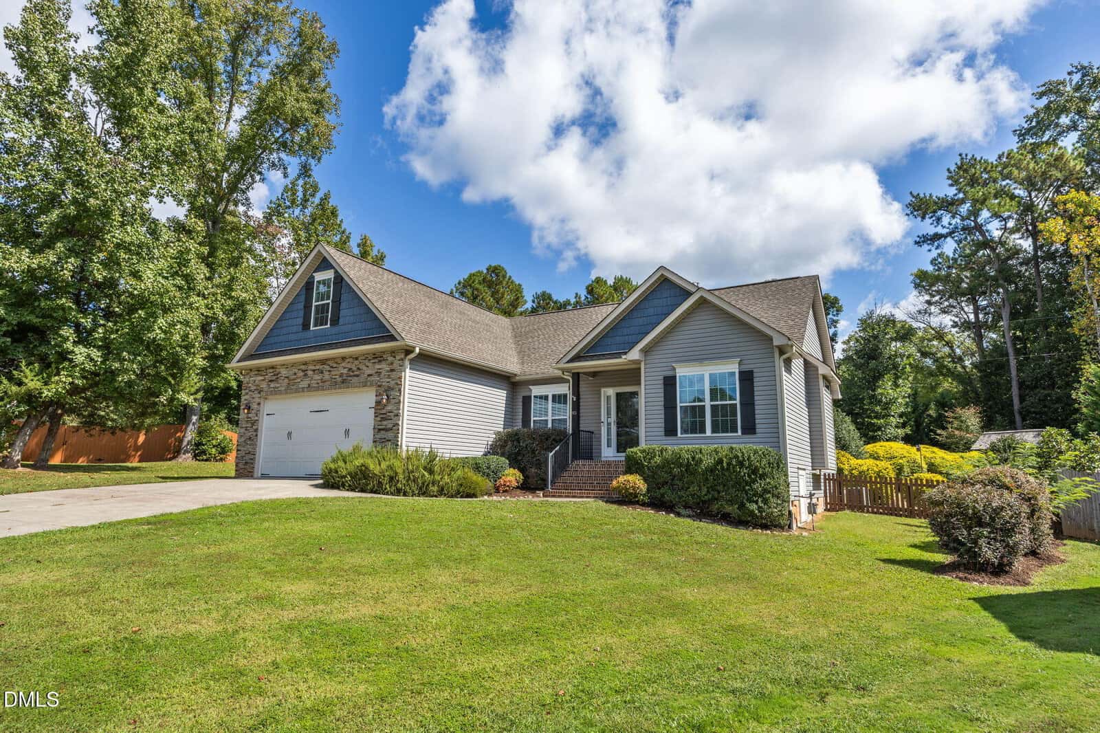 A single-story house with gray siding, a stone accent around the garage, a covered front porch, and landscaped yard, set on a sloped lawn with trees and a wooden fence in the background under a partly cloudy sky.