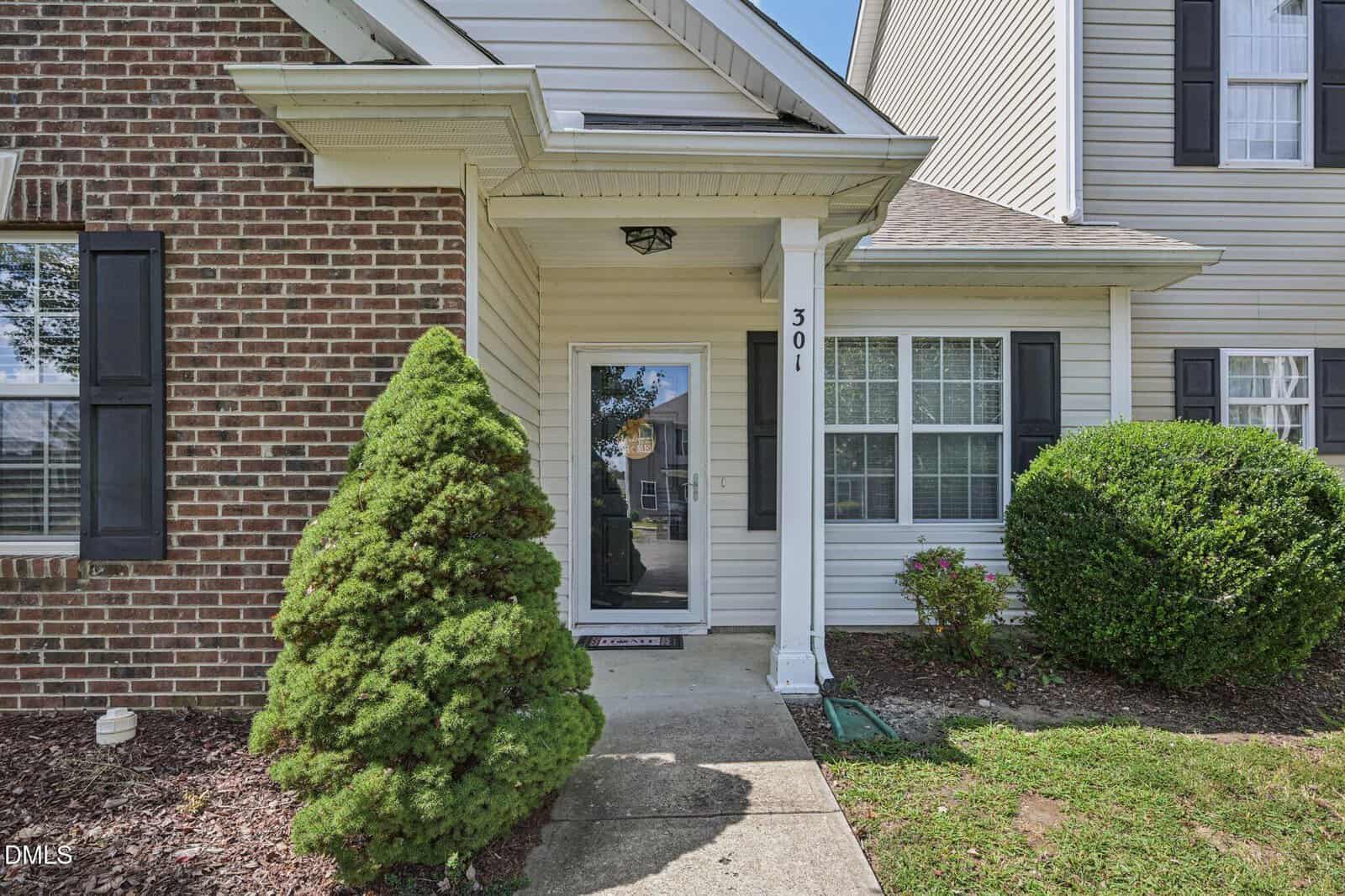 A front entrance of a house with a glass storm door, brick and siding exterior, windows with black shutters, two bushes, and a concrete walkway leading to the door, beneath a small covered porch.