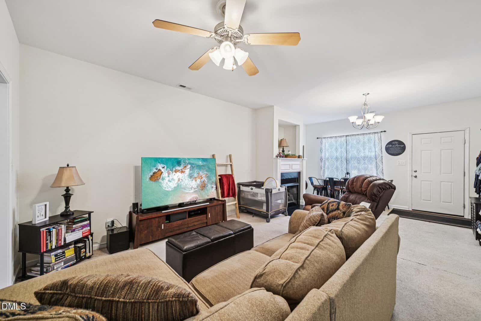 A living room with a beige sectional sofa, a TV displaying an aerial view of a beach, a ceiling fan with lights, a bookcase, a small fireplace, a baby crib, and a dining area with a table and chairs in the background.