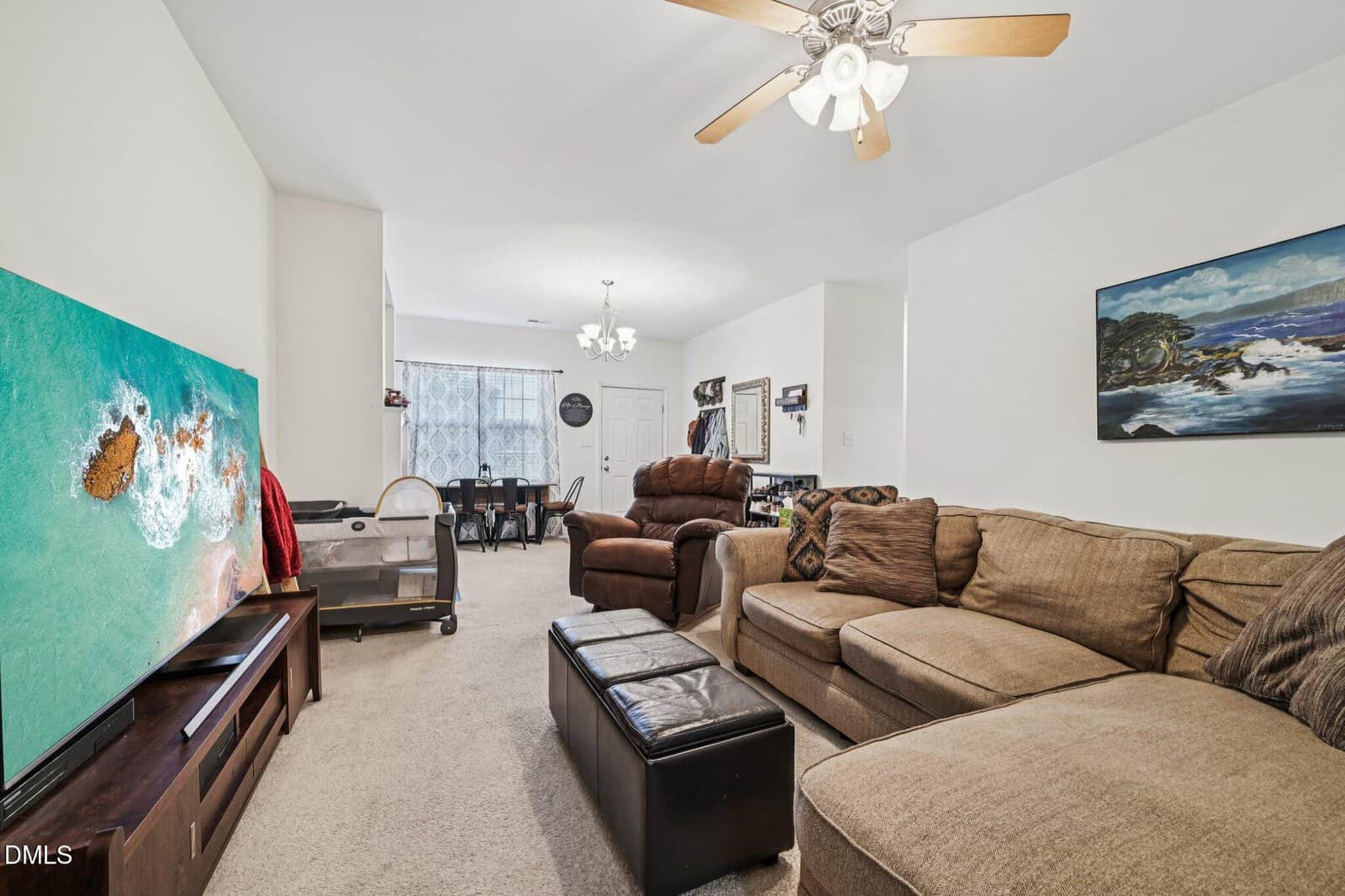 A living room with a beige sectional sofa, brown recliner, and ottoman, facing a large TV displaying an aerial beach scene. The space extends to a dining area with a table and chairs near a window.