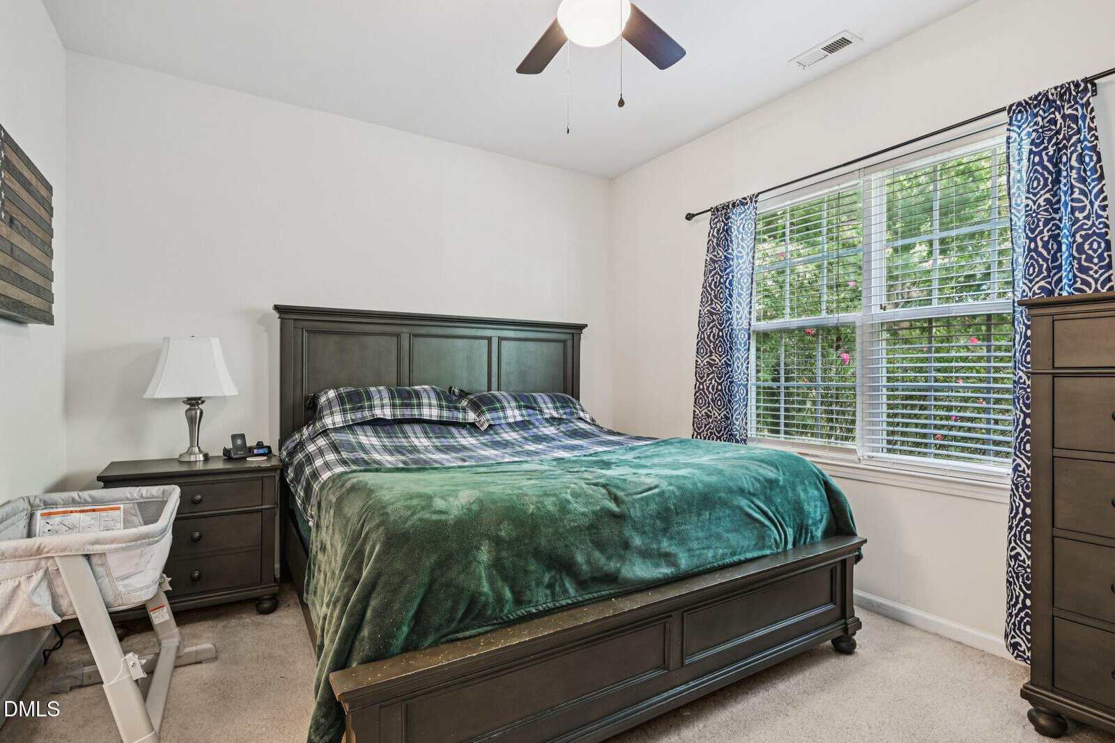 Bedroom with a dark wood bed, green and blue bedding, nightstand with lamp, bassinet, dresser, window with blue curtains, and ceiling fan. Natural light fills the room.