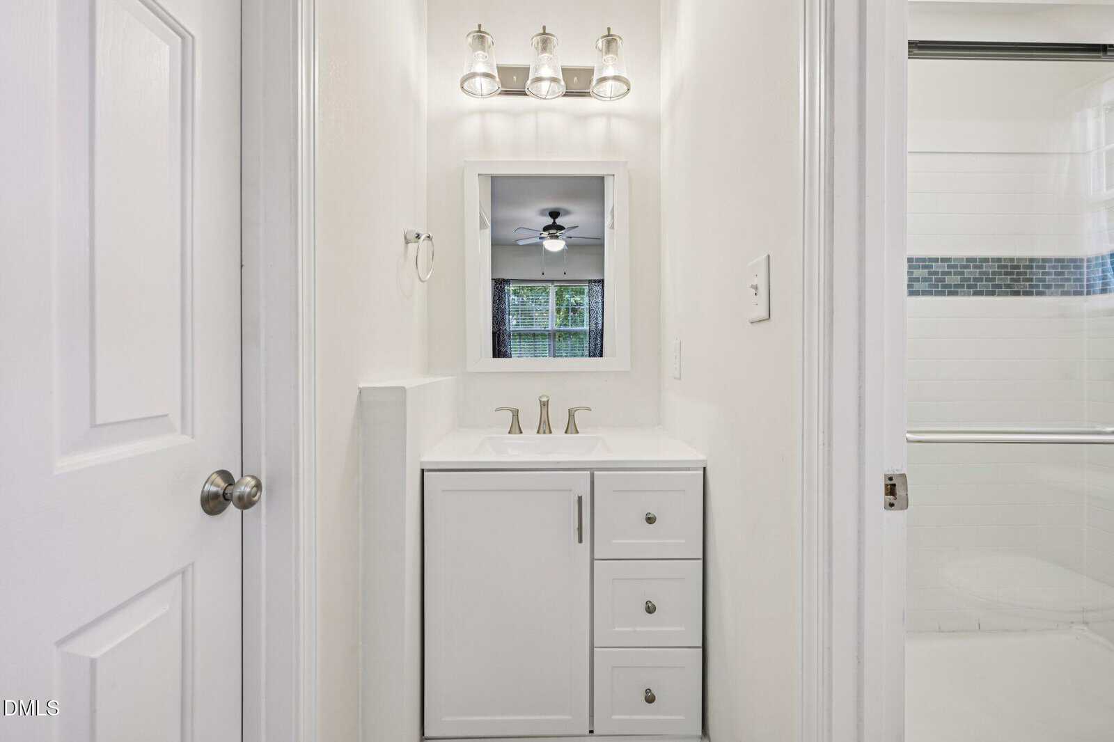Narrow white bathroom with a small vanity and mirror, three-light fixture above, and a glimpse of a shower with sliding glass door to the right. A ceiling fan and window are visible in the room beyond.