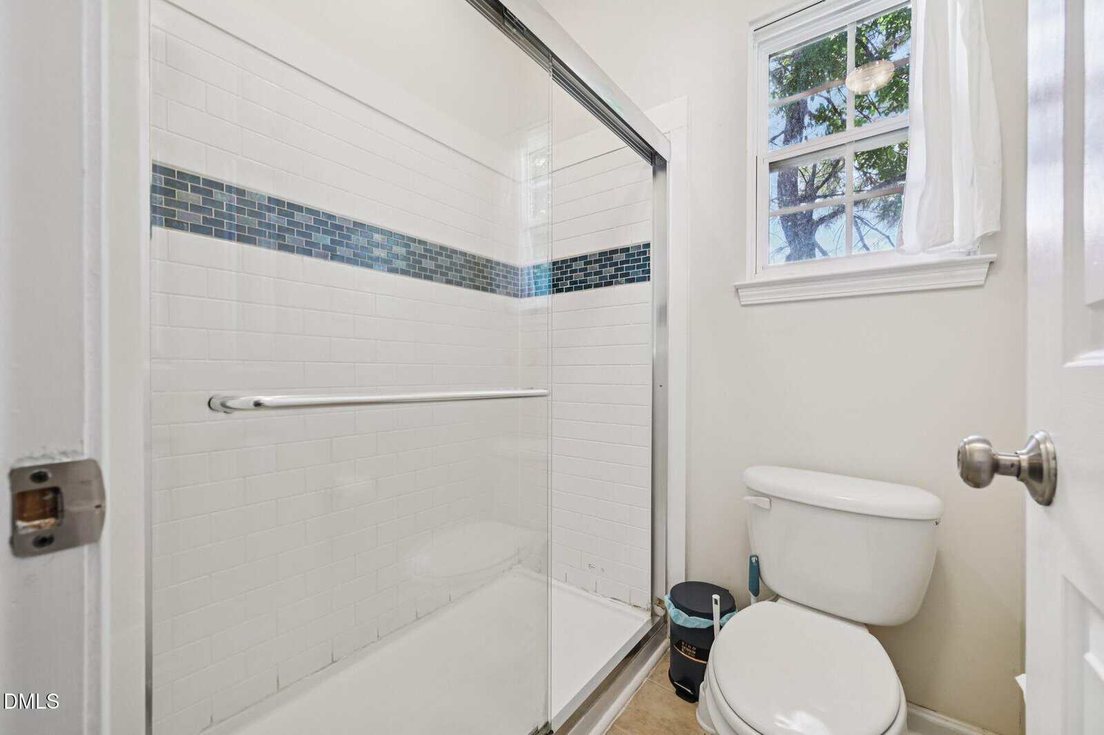 A bathroom with a glass-enclosed shower featuring white tiles and a blue accent strip, a toilet, a window with sheer curtains, and a small wastebasket next to the toilet.