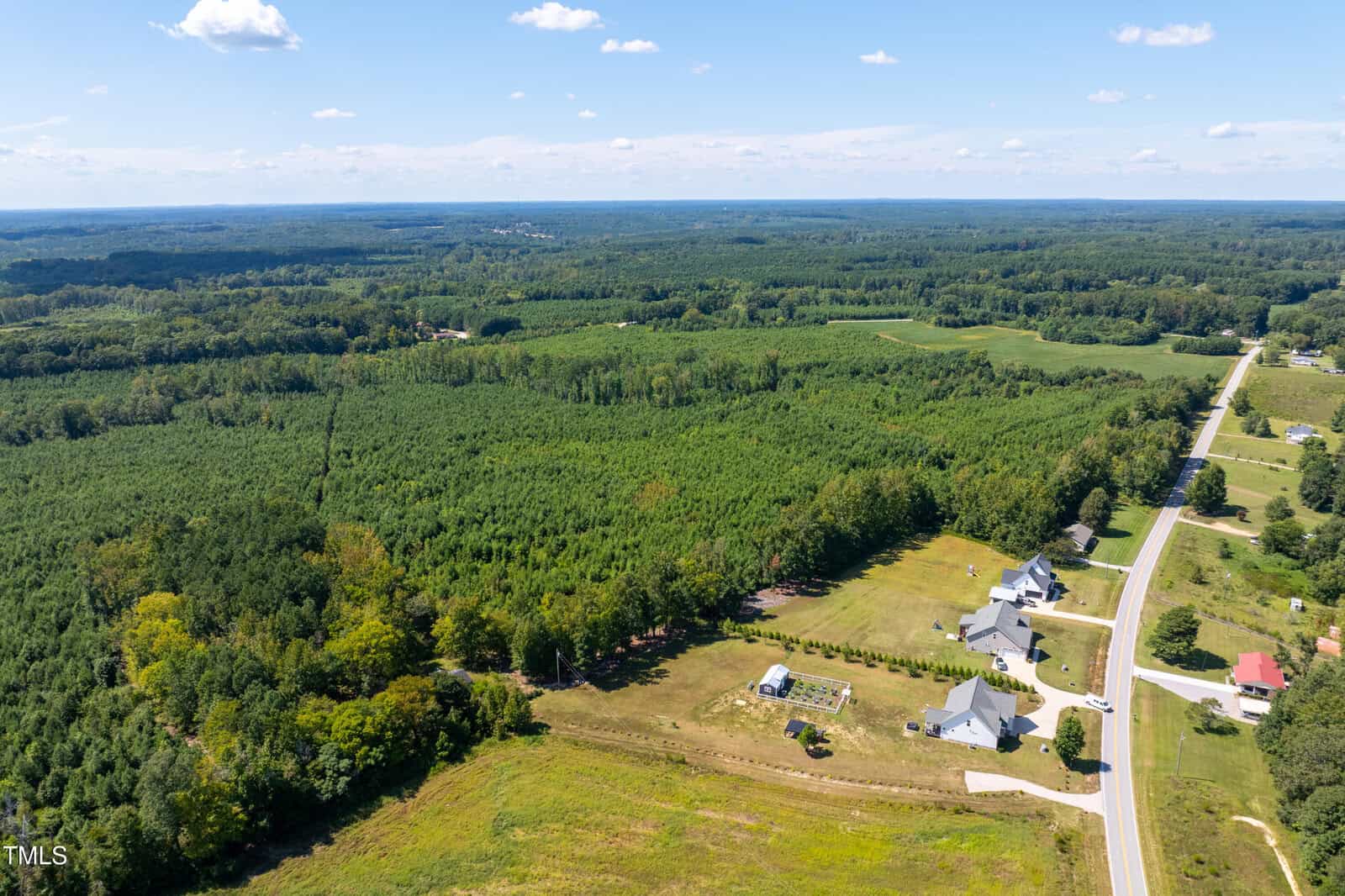 Aerial view of a rural area with dense forests, open fields, several houses, a fenced yard, and a long road running alongside the property under a clear blue sky.