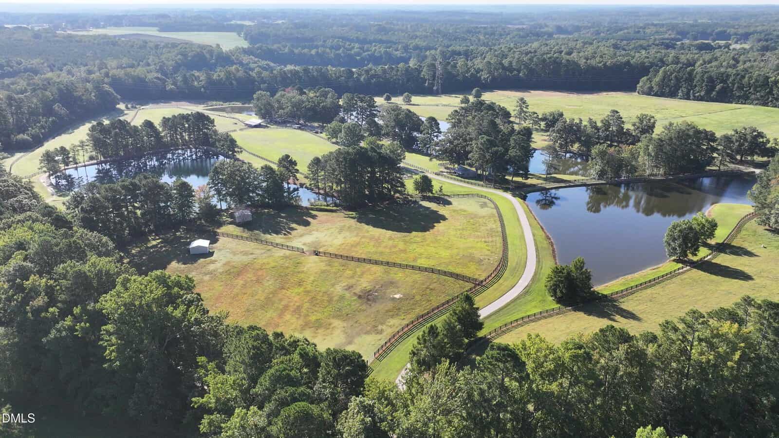 Aerial view of a rural landscape near Louisburg with fenced green fields, winding roads, several ponds, scattered trees, and a small white structure near the center. Dense forest borders the open land on multiple sides under clear skies, showcasing the serene setting of 6964 Pearces Road.