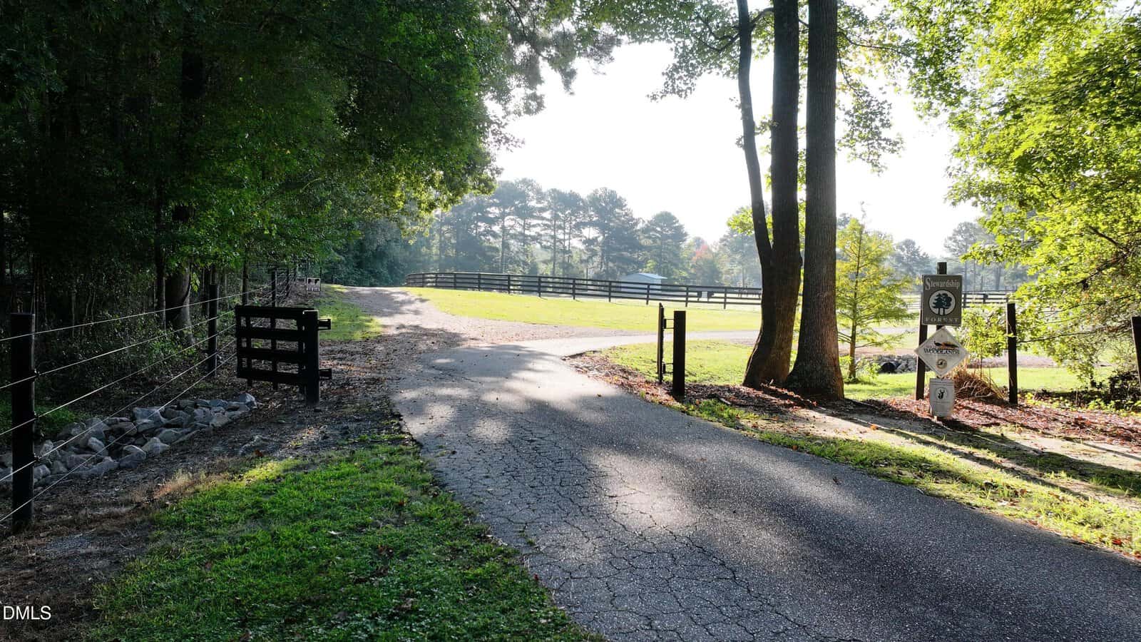 A paved driveway curves past a black gate and sign into a fenced, grassy area. Tall trees border the road, and sunlight filters through the branches. Fence lines and woods are visible in the background.