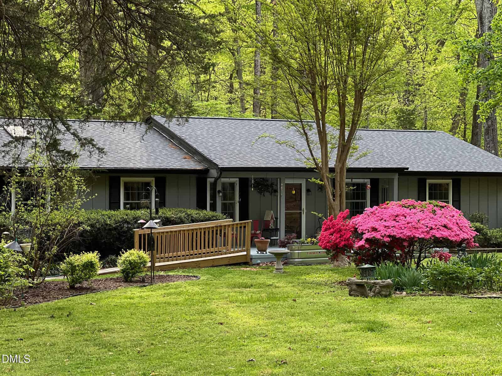 A single-story house with gray siding and a dark roof is surrounded by green grass, trees, and shrubs at 220 Cardinal Drive in Youngsville. A wooden ramp leads to the porch, and pink azalea bushes bloom in the garden near the walkway.