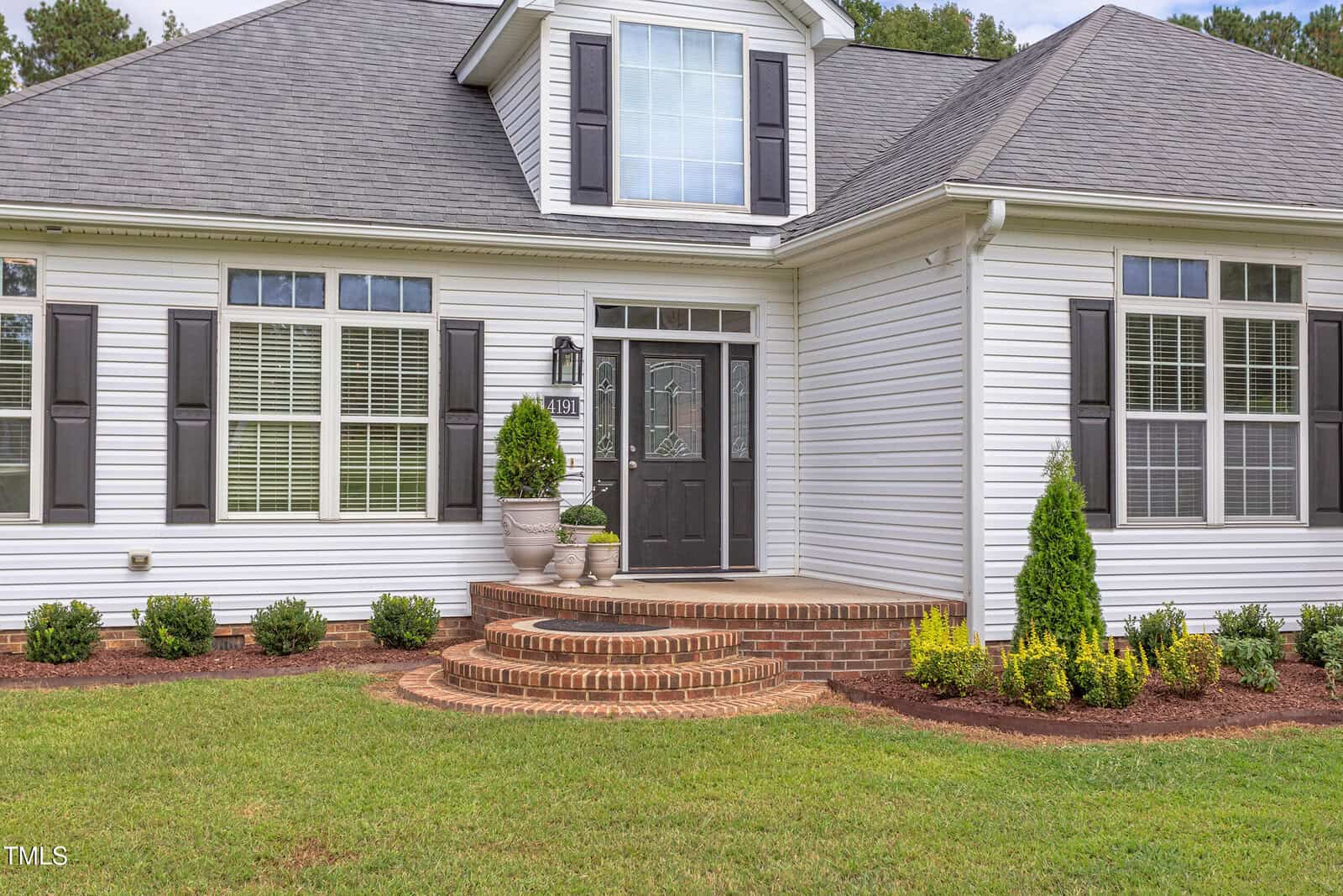 A white house with gray shutters and a gray front door, featuring a small porch with brick steps, potted plants, trimmed bushes, and a well-maintained lawn in front.