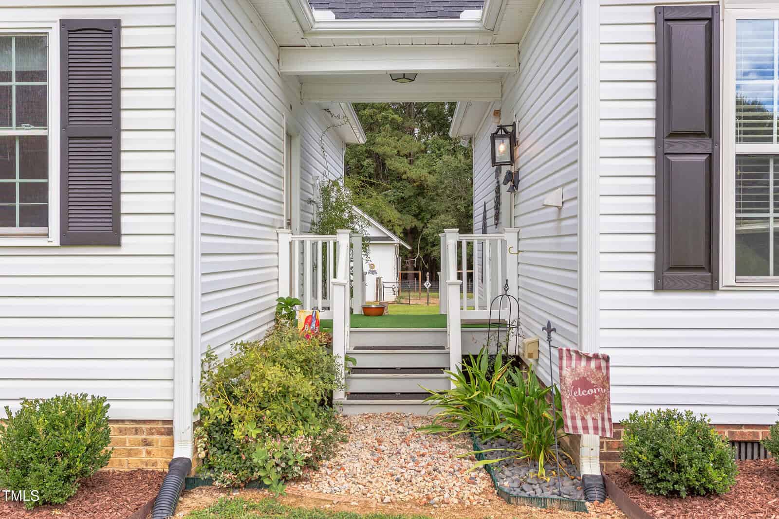 A gravel walkway leads to a small porch with steps between two white-sided sections of a house. There are green plants, a decorative flag, and a lantern near the entrance. A yard and another building are visible in the background.