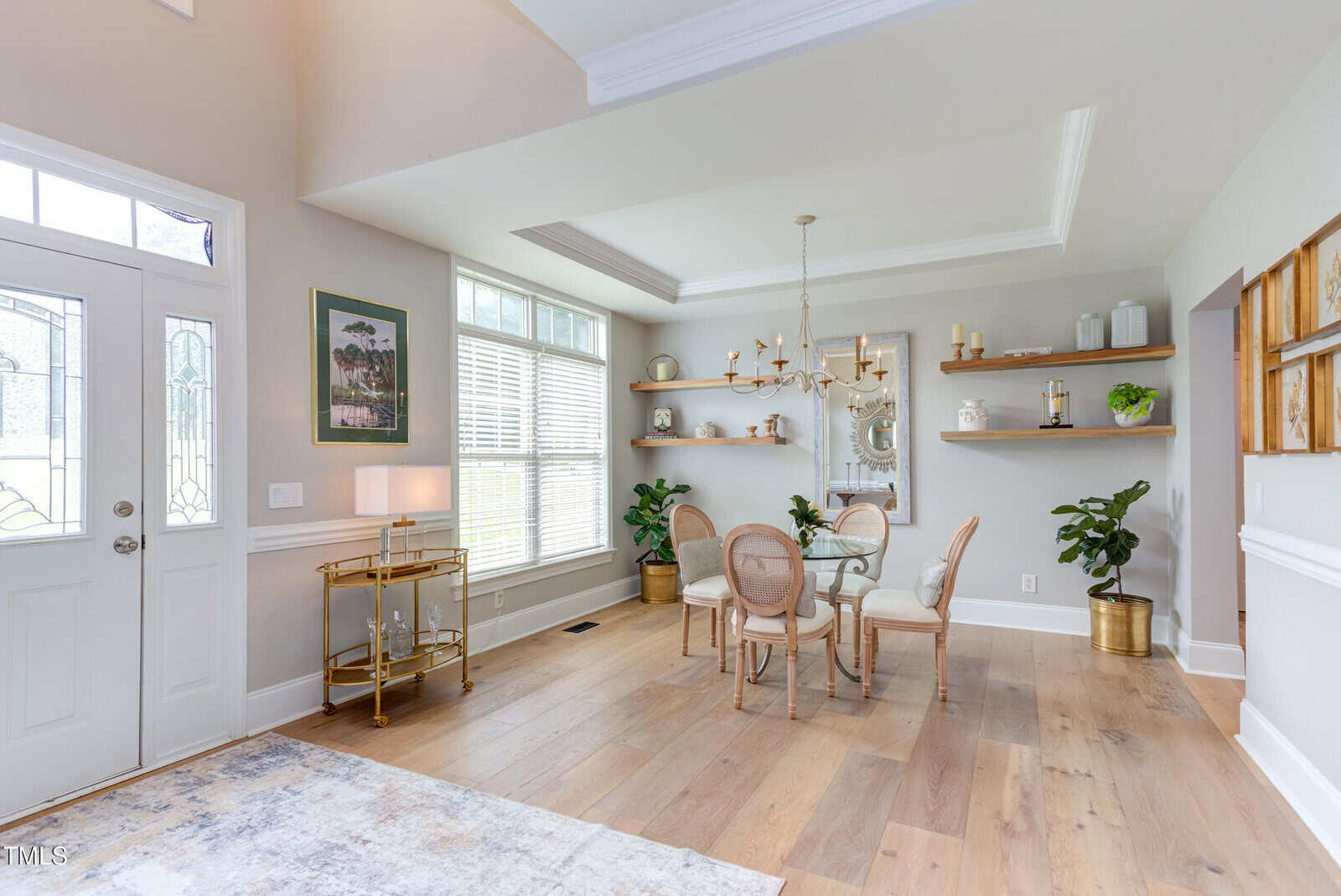 A bright dining room with a glass table, four chairs, gold chandelier, and two potted plants. Large windows and light wood floors add to the airy feel. Shelves with decor items are on the wall. A bar cart is near the door.