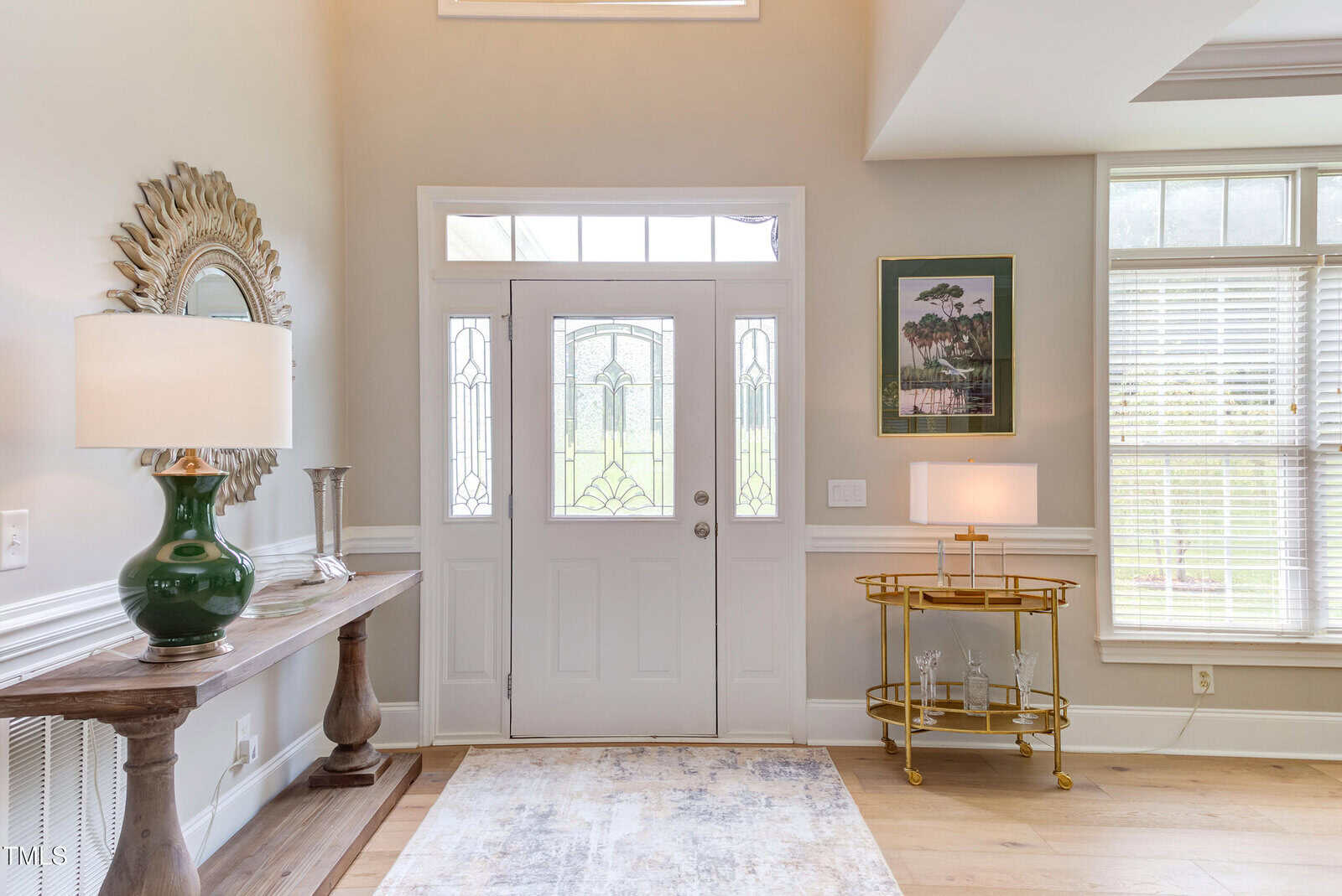 A bright entryway with a white front door, sidelights, a wooden console table with a lamp and mirror on the left, a gold bar cart with a lamp and framed art on the right, and a light-colored rug on the floor.