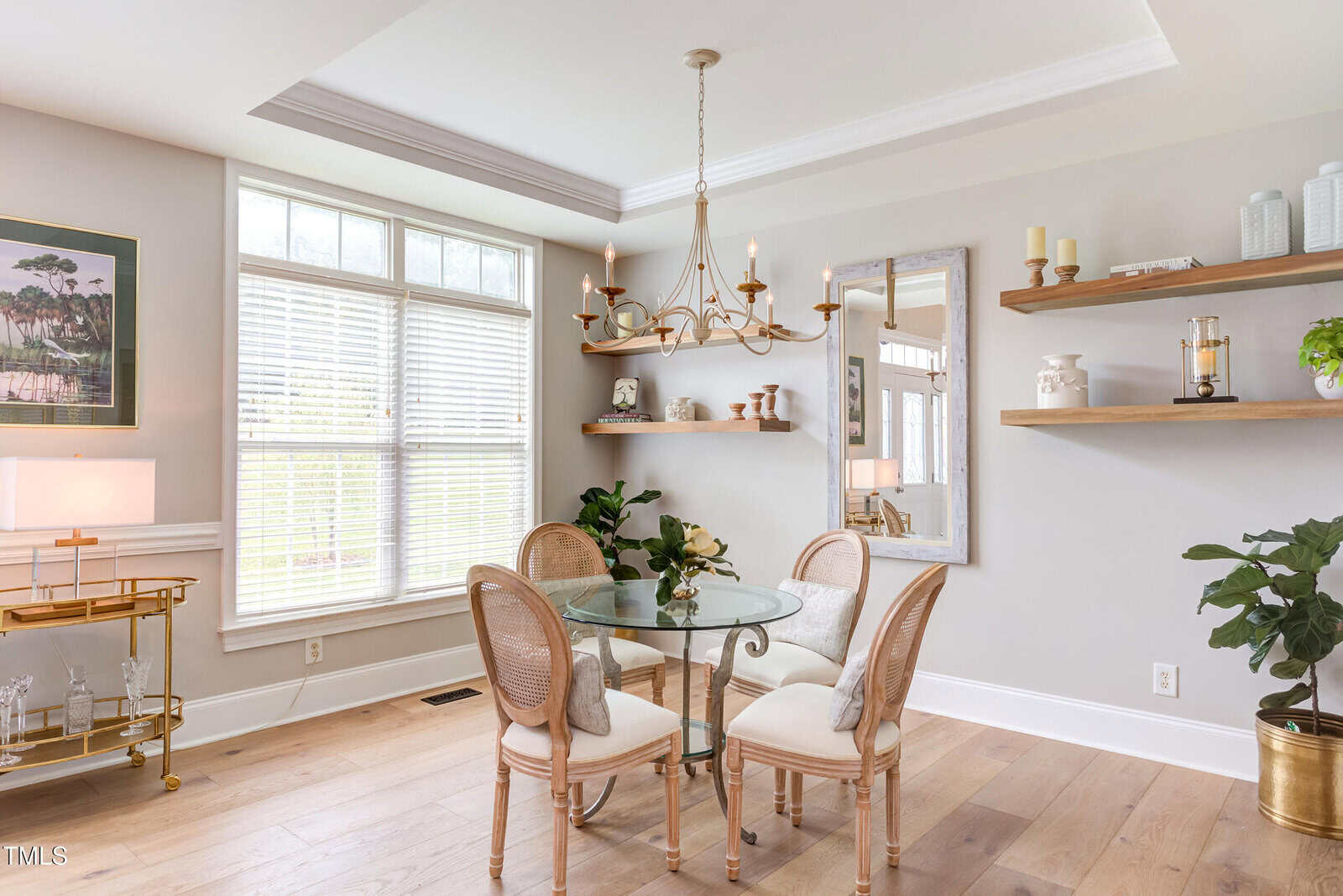 A dining room with a glass table and four chairs on light wood flooring. A gold chandelier hangs above, and the walls have shelves with decor, a mirror, and a large window letting in natural light.