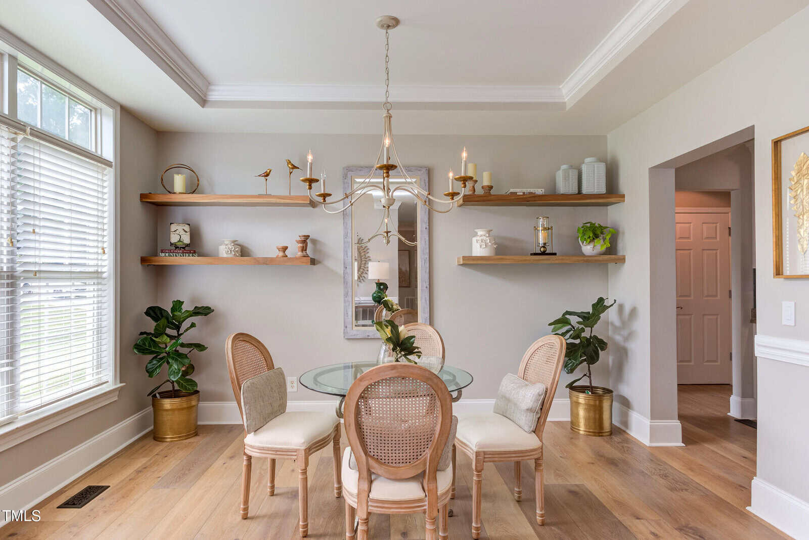 A dining room with a round glass table and four chairs. Wooden shelves with decorative items line the back wall. Two potted plants stand in gold planters. A chandelier hangs from the ceiling, and a window lets in natural light.