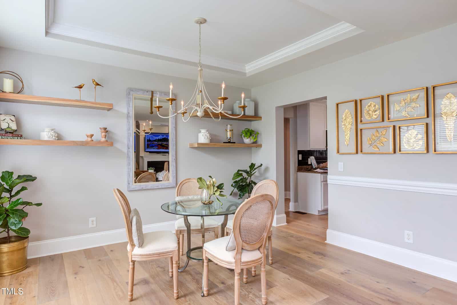 Bright dining room with a glass round table, four beige chairs, gold chandelier, wall art of gold leaves, floating wood shelves with decor, and potted plants on light wood flooring.