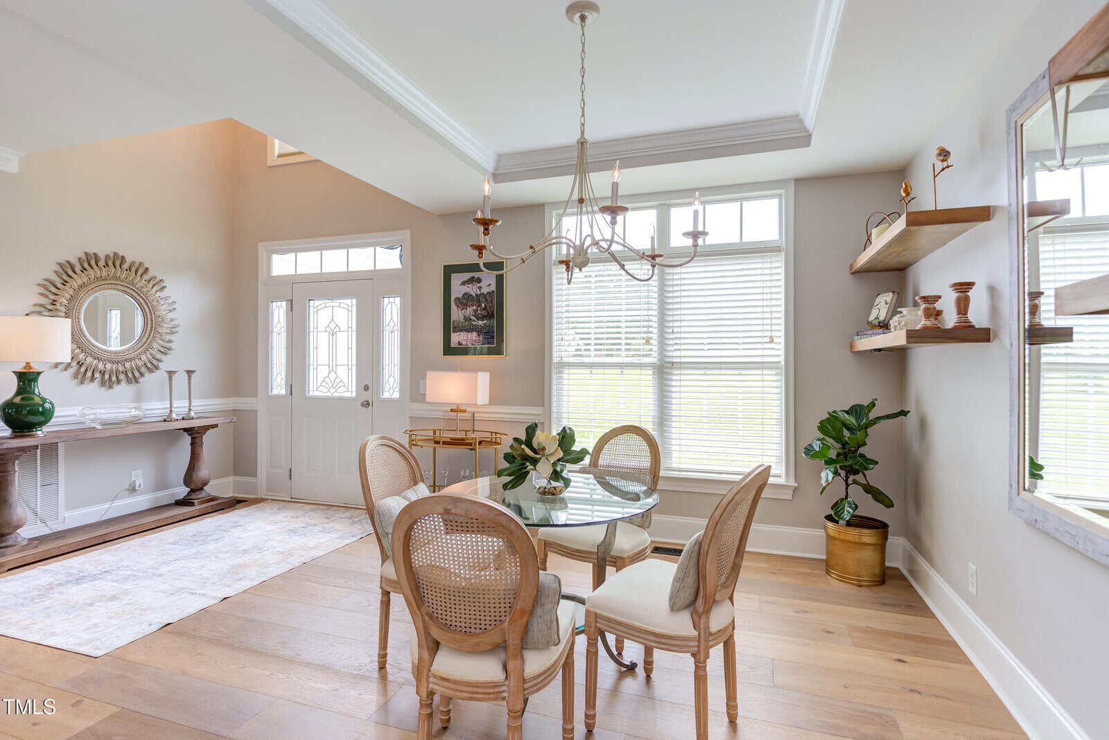 Bright dining area with a round glass table, four wicker chairs, chandelier, large windows, wooden shelves with decor, potted plant, and an entryway with a mirror, console table, and decorative items.