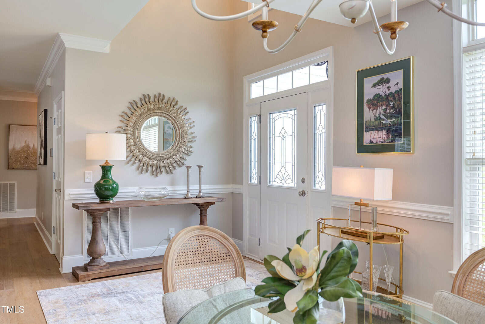 A bright entryway with a glass front door, a wooden console table with a lamp and mirror, a gold bar cart with a lamp, and a round glass dining table with a flower arrangement in the foreground.