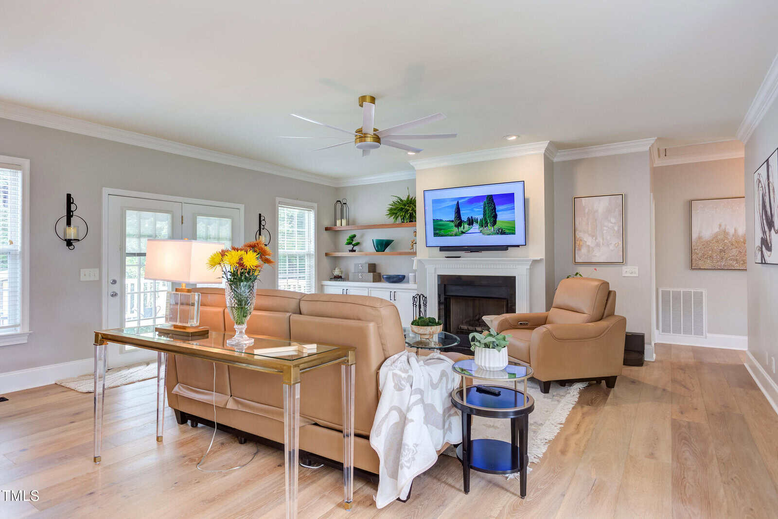 Bright living room with tan leather sofas, a glass table with flowers, modern decor, wall-mounted TV above a fireplace, light wood flooring, and large windows allowing natural light.