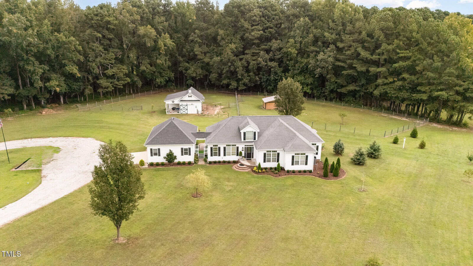 A single-story white house with a gray roof sits on a large grassy property with a circular gravel driveway, surrounded by trees. Two outbuildings are visible in the background near a wooded area.