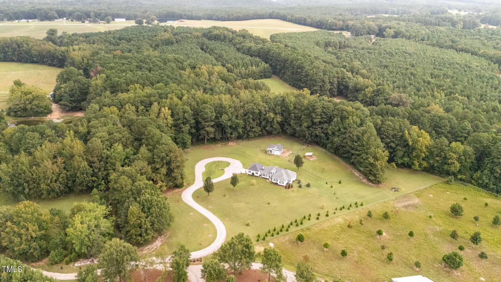 Aerial view of a large house with a circular driveway, surrounded by trees and open fields, located in a rural area with dense forest in the background.