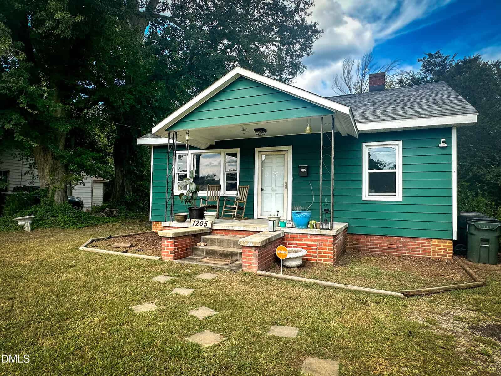 A small green house with white trim and a covered front porch, set on a grassy yard with stepping stones leading to the steps. There are potted plants and a rocking chair on the porch, with trees and a blue sky in the background.