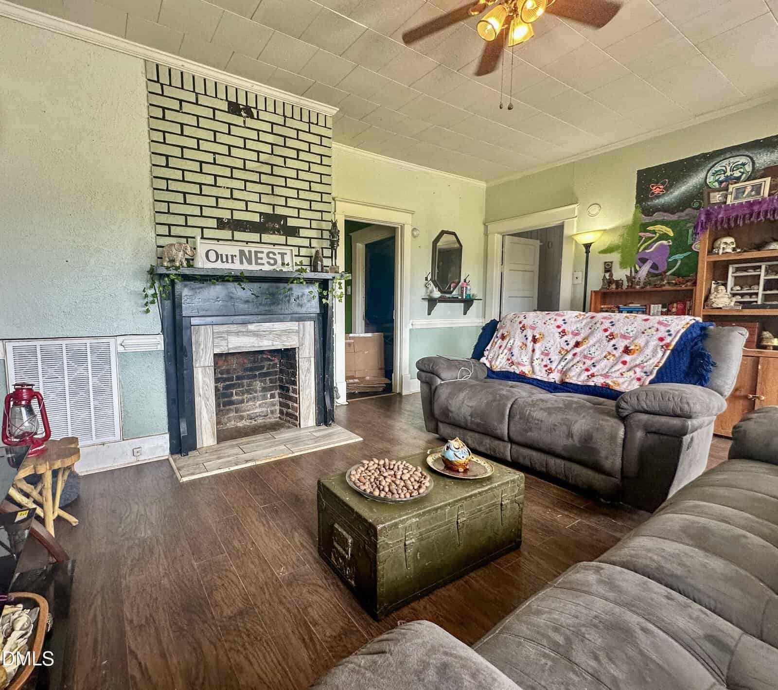 A cozy living room with two gray sofas, a brick fireplace with a sign reading “Our NEST,” wooden floors, a green trunk as a coffee table, and various decor items on shelves and walls. Ceiling fan above.