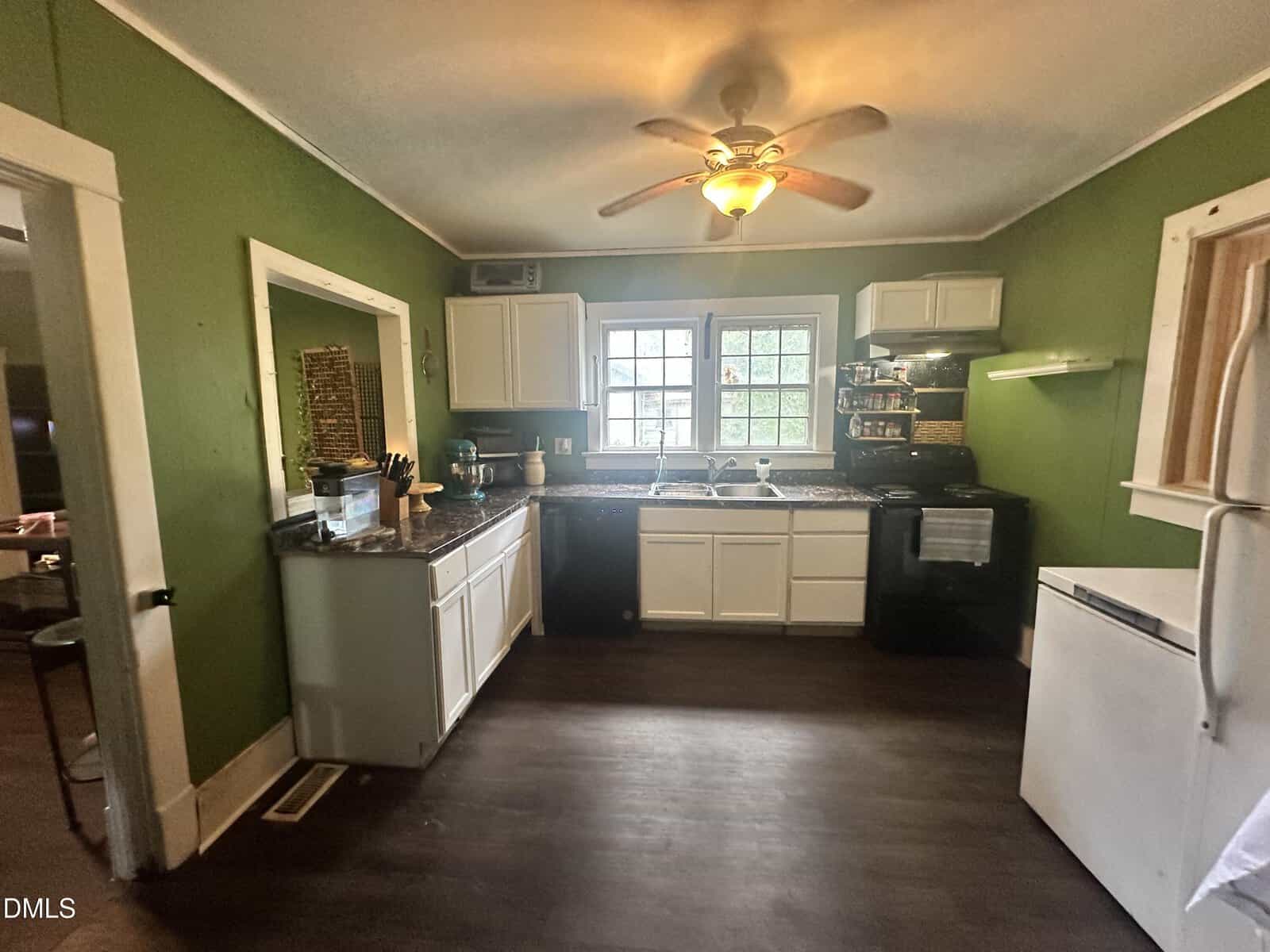 A kitchen with green walls, white cabinets, dark countertops, a ceiling fan with a light, a white refrigerator, a black stove, a window above the sink, and dark wood floors.