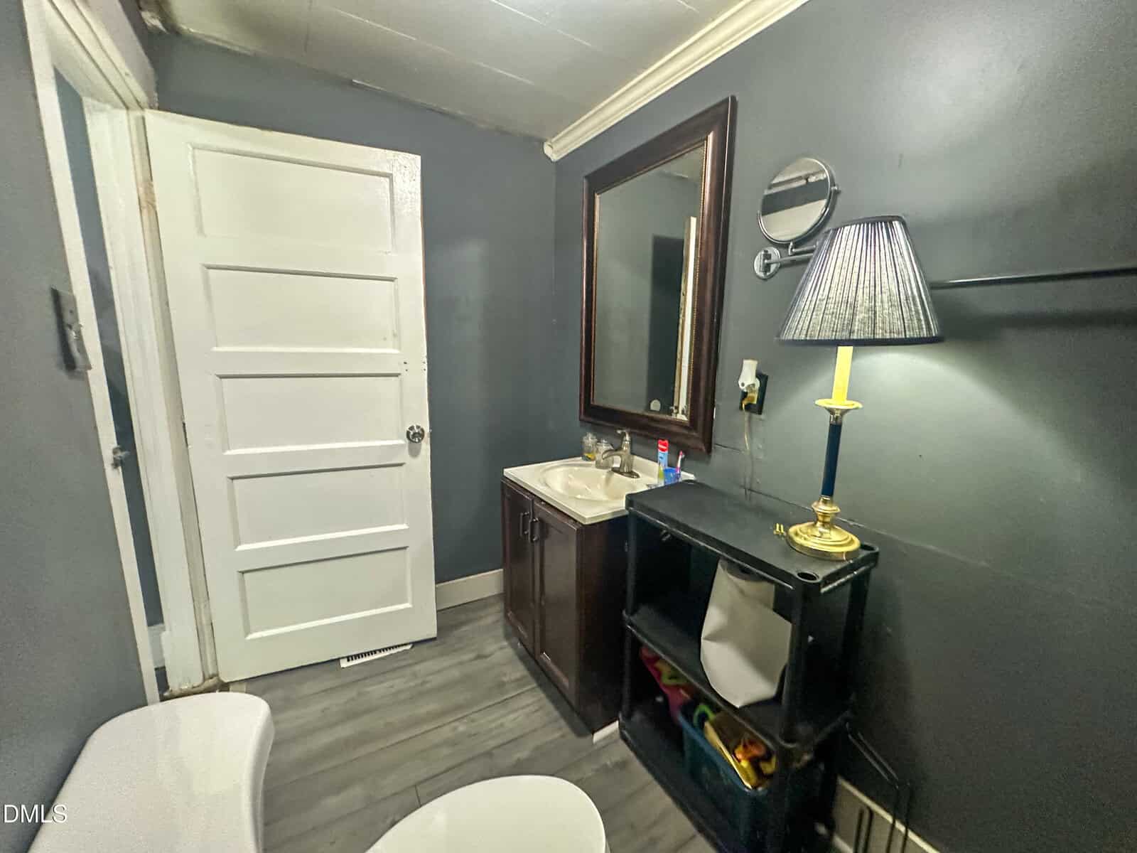 A small bathroom with dark gray walls, a white door, a sink with a mirror above it, a black shelf with a lamp, toiletries, and a white toilet partially visible in the foreground.