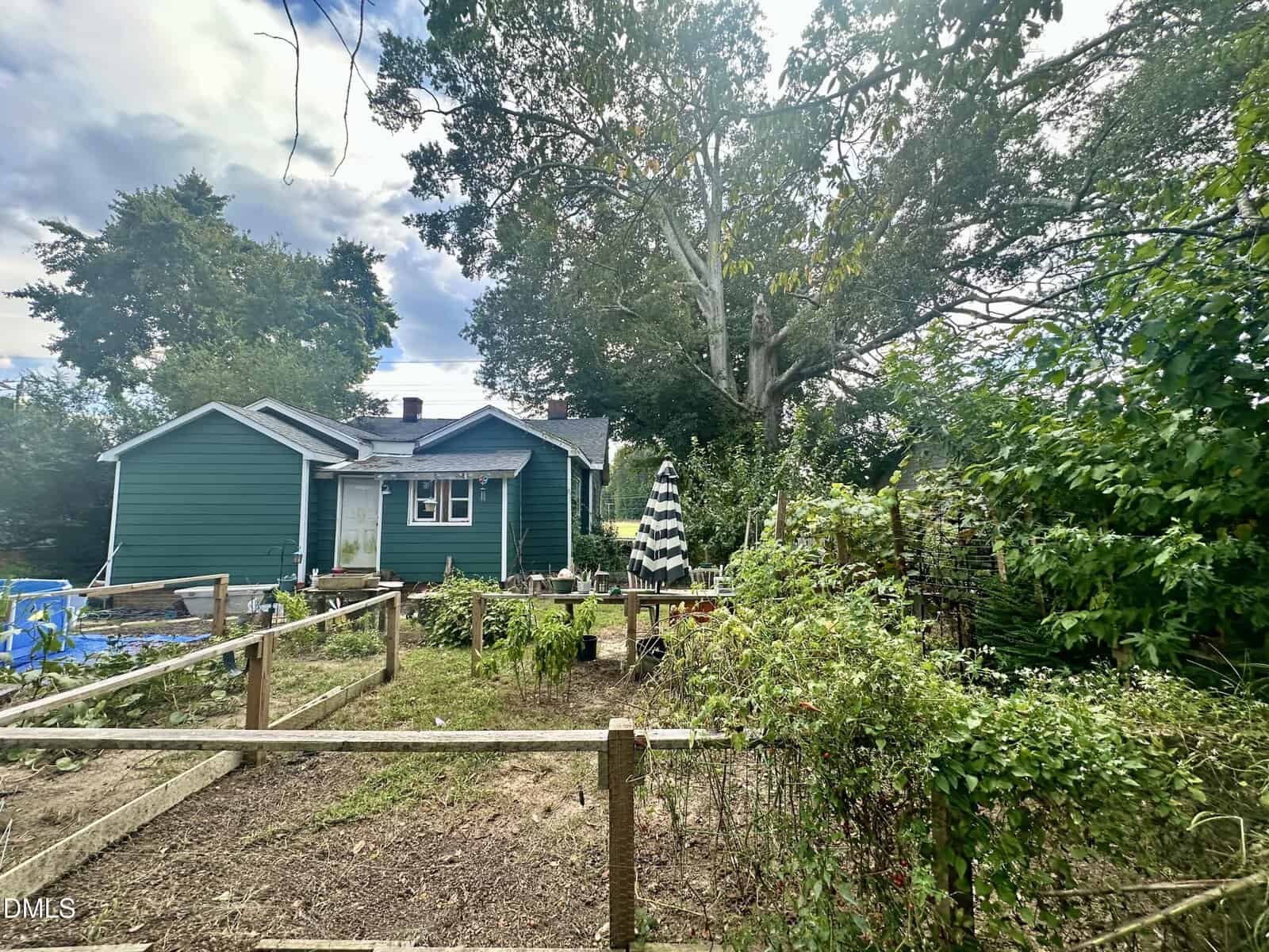 A green house with white trim sits behind a garden with raised beds and overgrown plants. A black-and-white striped umbrella shades a picnic table next to the house, and large trees surround the yard under a partly cloudy sky.