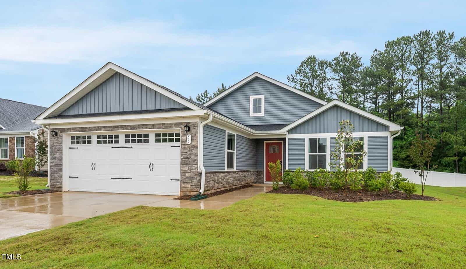 Charming and inviting, this single-story house at 55 Boots Ridge Way in Franklinton features blue-gray siding, stone accents, a double garage, and a striking red front door. The welcoming front porch overlooks a well-kept lawn, while trees and shrubs line the walkway leading to the entrance. A peaceful wooded area provides a scenic backdrop beneath an overcast sky.