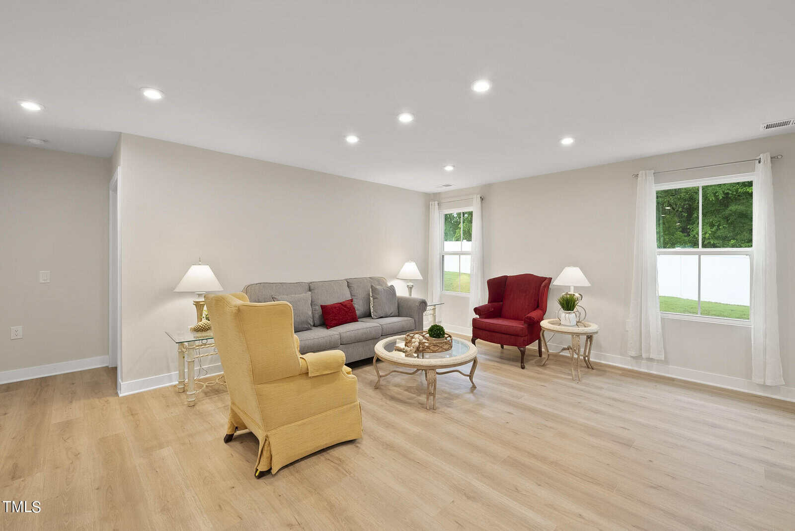 A living room with a gray sofa, red and yellow armchairs, glass coffee tables, lamps, wood flooring, and two windows with white curtains showing a green yard outside. Walls are light-colored with recessed ceiling lights.
