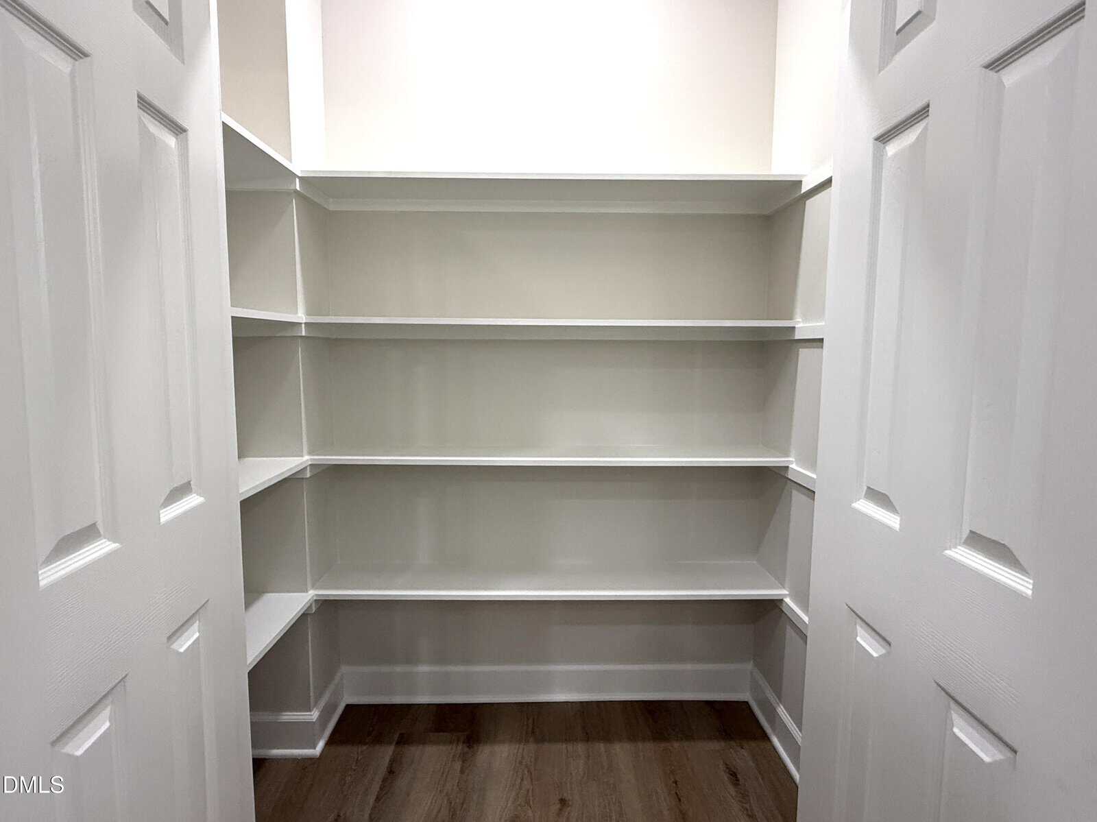 A walk-in pantry with white built-in shelves on three walls, double white doors, and a wood floor. The shelves are empty and the space is well-lit.