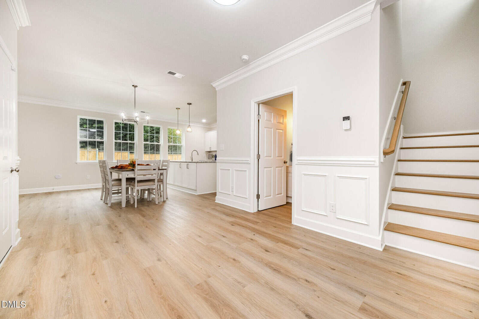 A bright, modern open-concept dining and kitchen area with light wood floors, white walls, a dining table with chairs, recessed lighting, and a staircase leading upstairs.