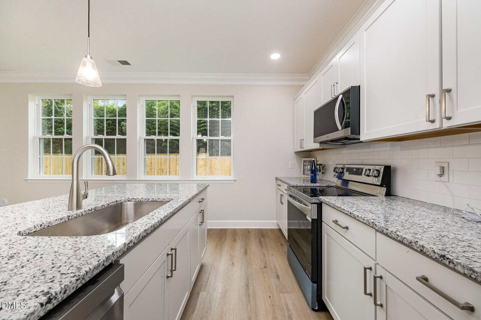 A modern kitchen with white cabinets, granite countertops, stainless steel appliances, a center island with a sink, pendant lighting, and large windows letting in natural light.