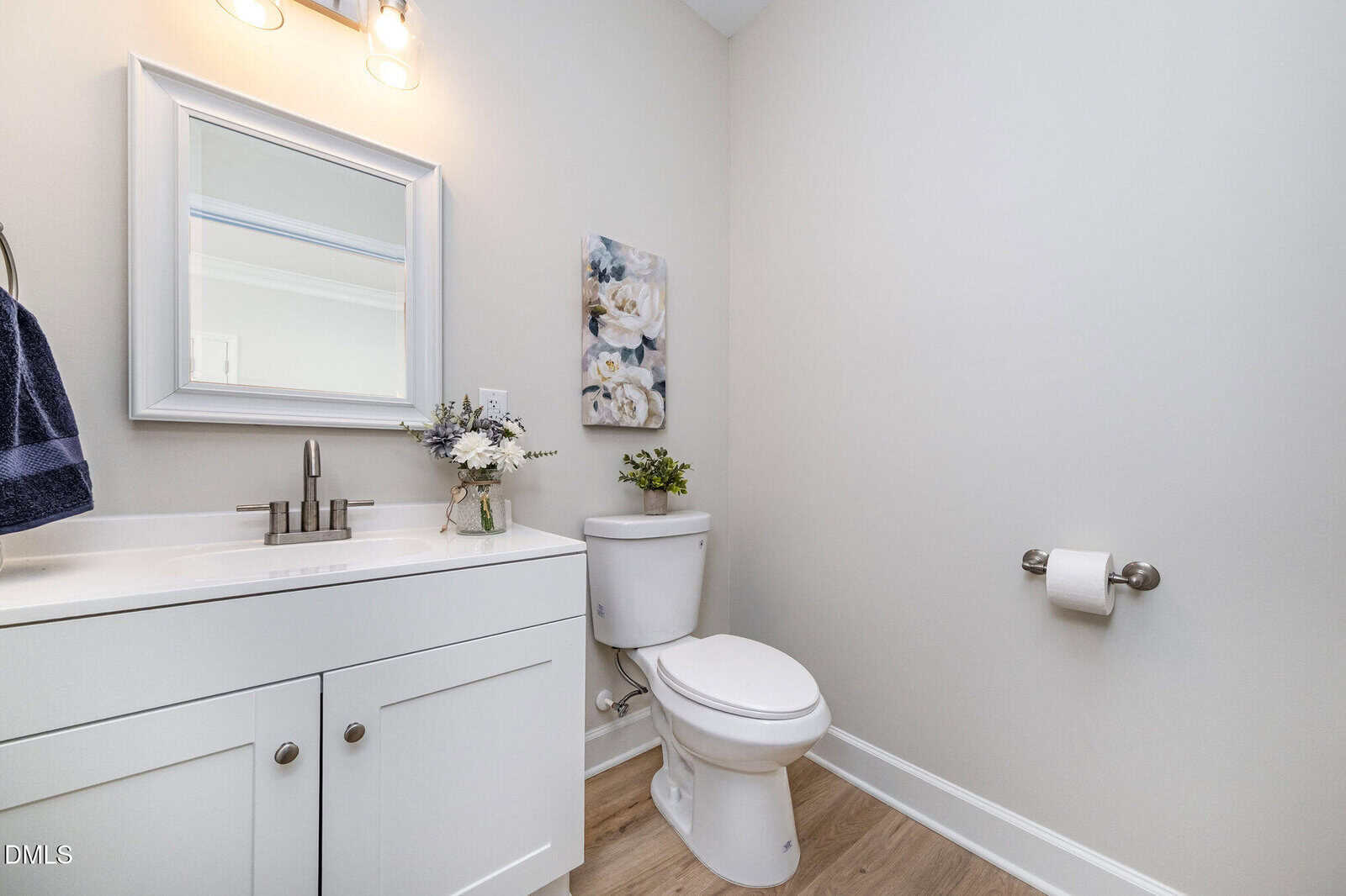 Modern bathroom with white vanity, sink, and mirror on the left, toilet and floral wall art on the right, wooden floor, light walls, and a towel and toilet paper holder.