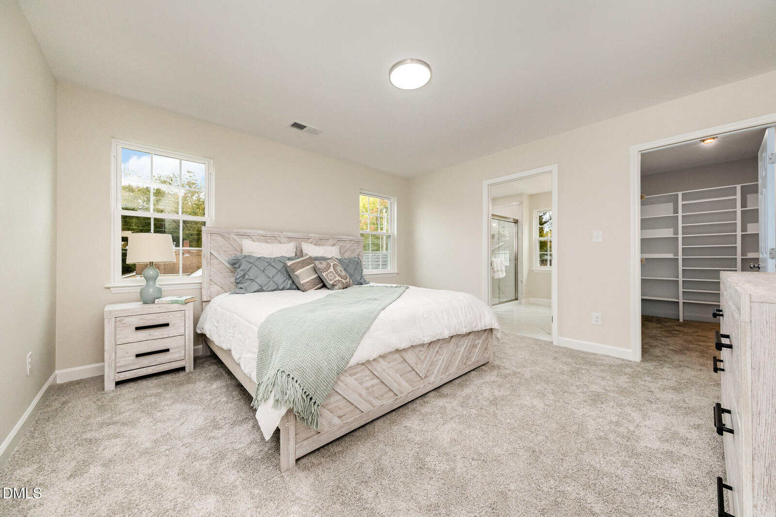Bright bedroom with a large bed, nightstand, two windows, carpeted floor, and a neatly organized walk-in closet visible through an open door. A bathroom with glass shower doors is also seen. Walls are painted light beige.