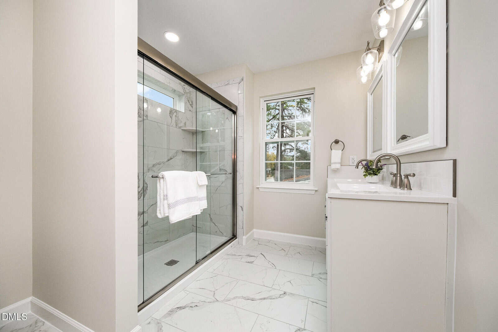 Modern bathroom with large glass shower, white tile floor, single vanity with sink and mirror, light fixtures above, and a window letting in natural light. White and neutral tones create a clean, bright look.